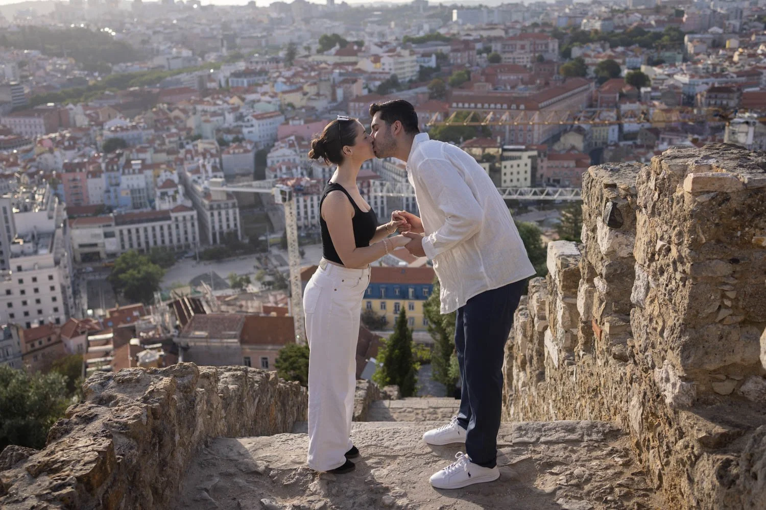A couple sharing a kiss on a stone ledge with a cityscape in the background.