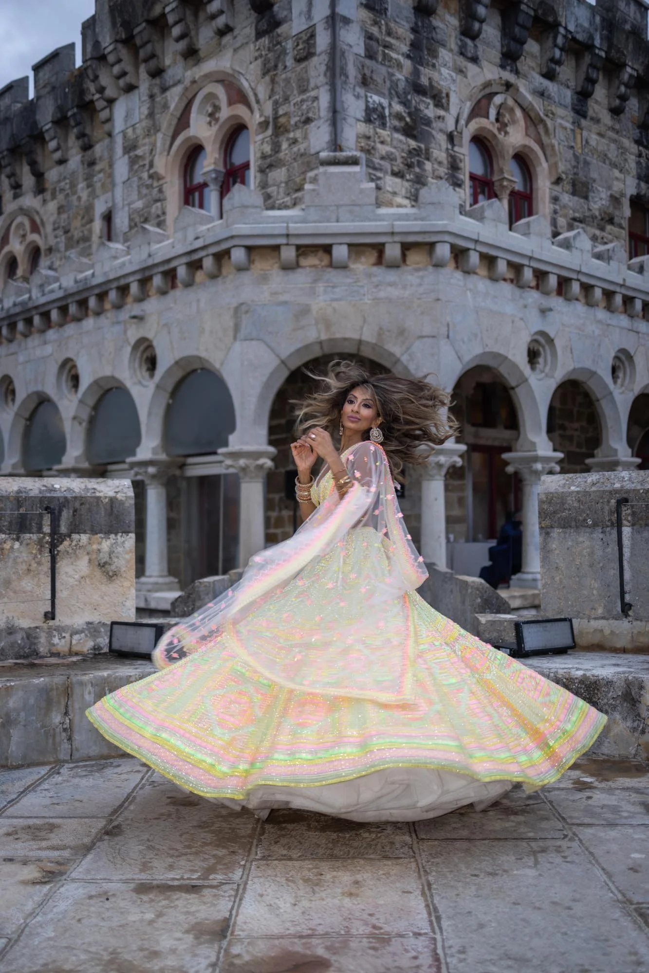 A woman in a colorful, flowing dress twirls around in front of a historic stone building with arched windows and decorative architecture.