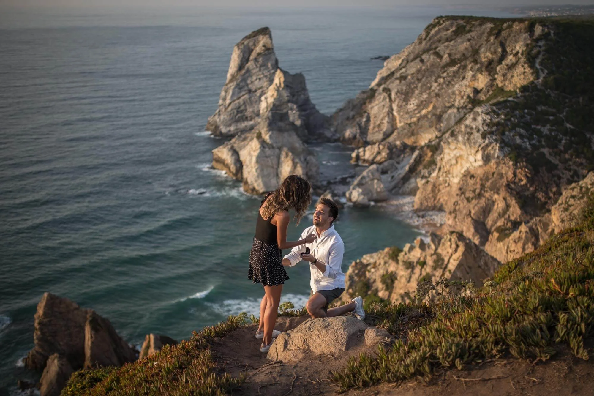 A man proposing marriage to a woman on a rocky cliffside overlooking the ocean at sunset with large rocky formations in the background.