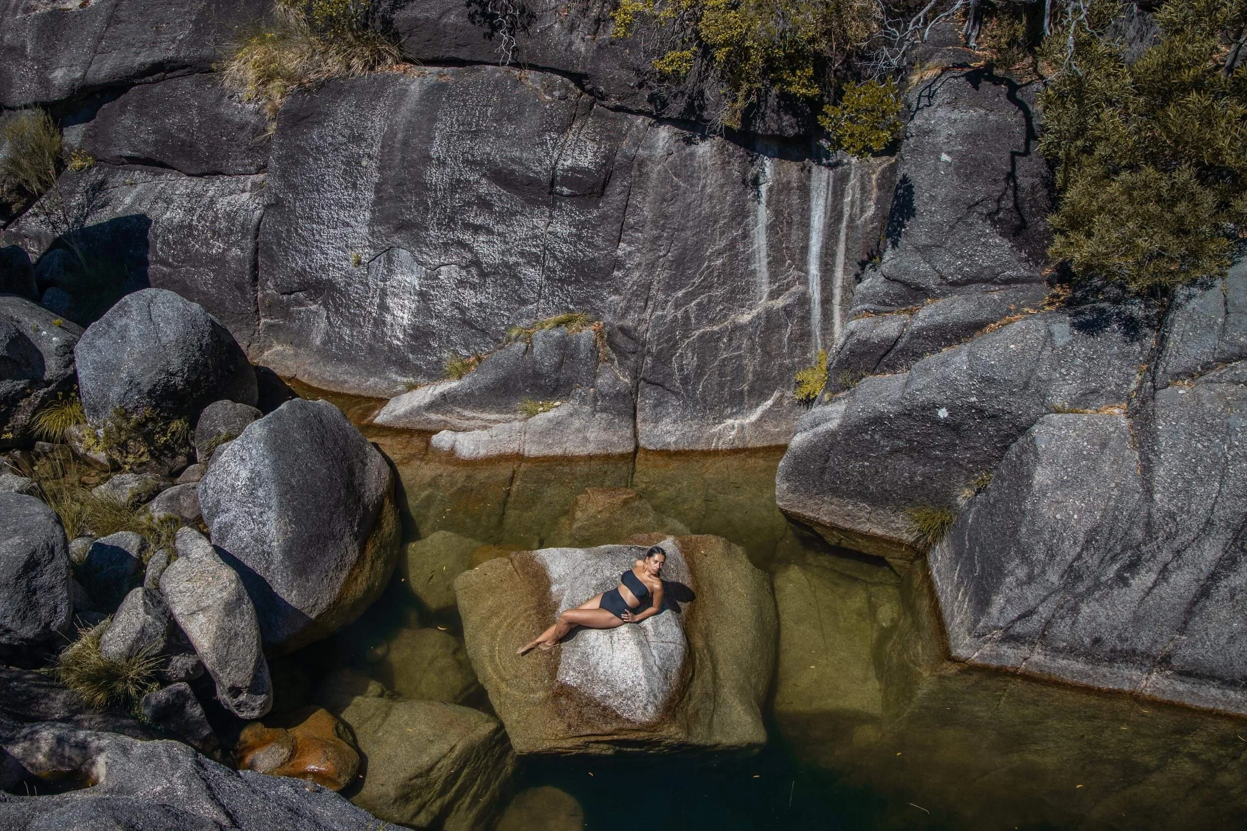 A woman in a black swimsuit lying on a large rock in a small pool of water surrounded by large gray rocks and boulders, with a waterfall cascading down a rocky cliff in the background.