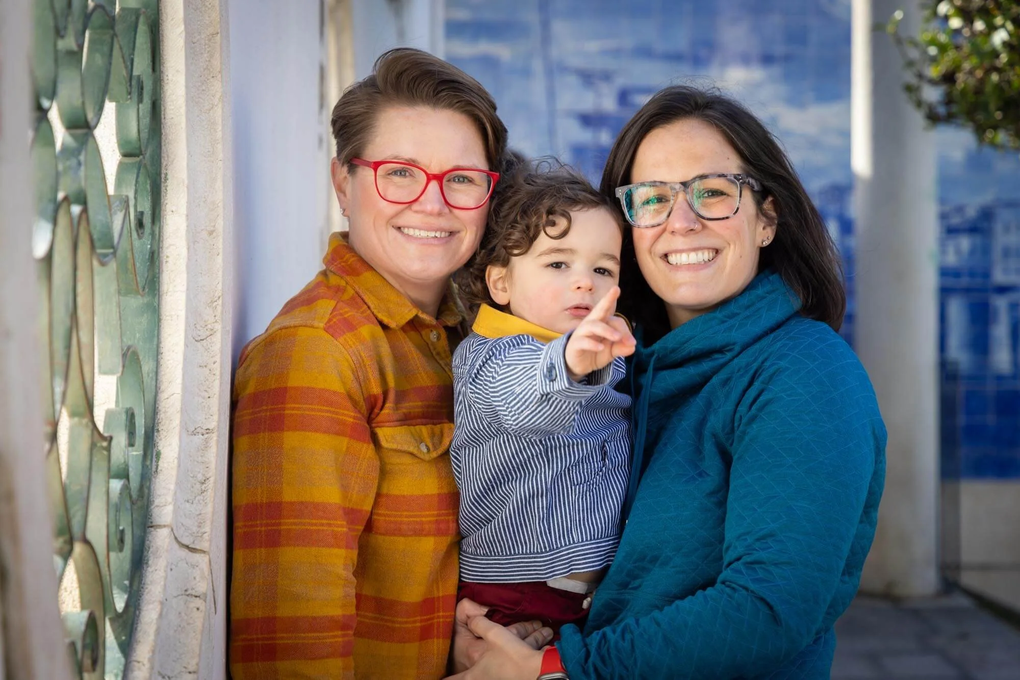 Two women and a young boy standing outside near a wall with green decorative ironwork, smiling at the camera. They are all posing for a photographer in Lisbon for their family photoshoot,