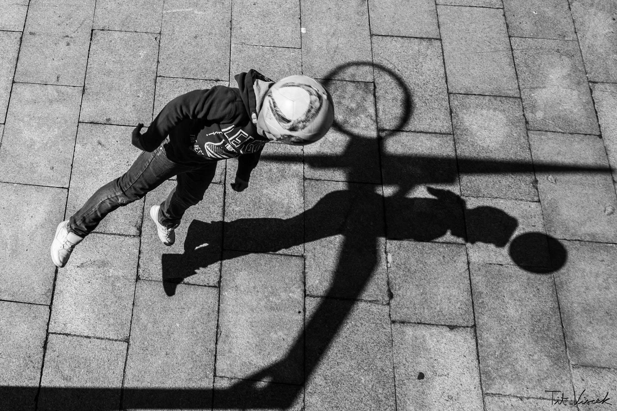 A young boy in a hoodie and sneakers playing basketball on a paved outdoor surface, with his shadow and the basketball's shadow cast on the ground.