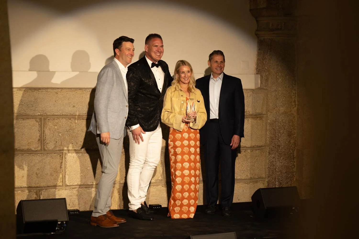 Four people standing together on a stage, three men and one woman, with a woman holding an award or trophy. They are dressed in formal and semi-formal attire and are smiling. The background consists of a stone wall and a projector speaker is visible 