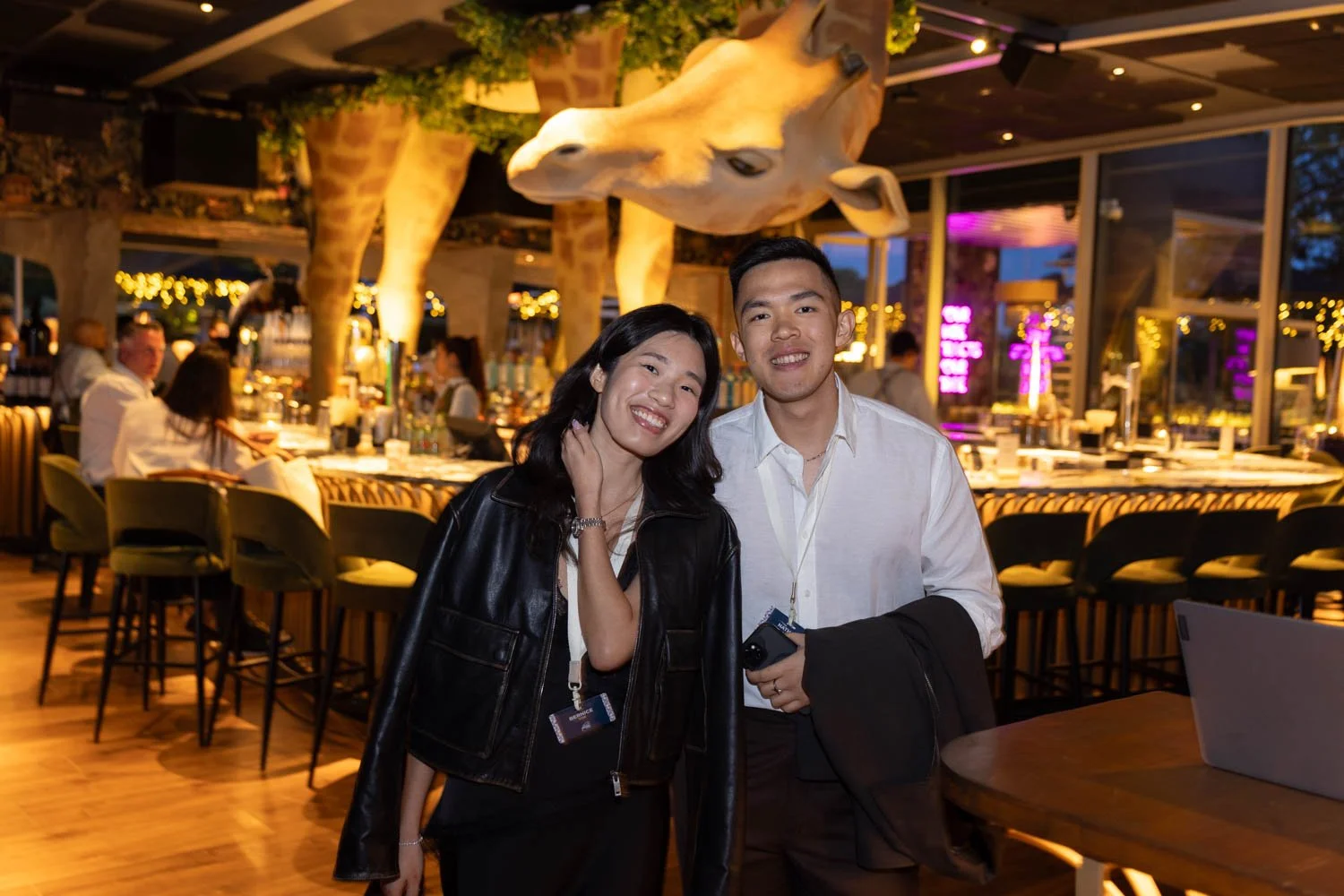 A woman and a man smiling at the camera in a restaurant in Lisbon with giraffe decorations and warm lighting, with tables and people in the background.