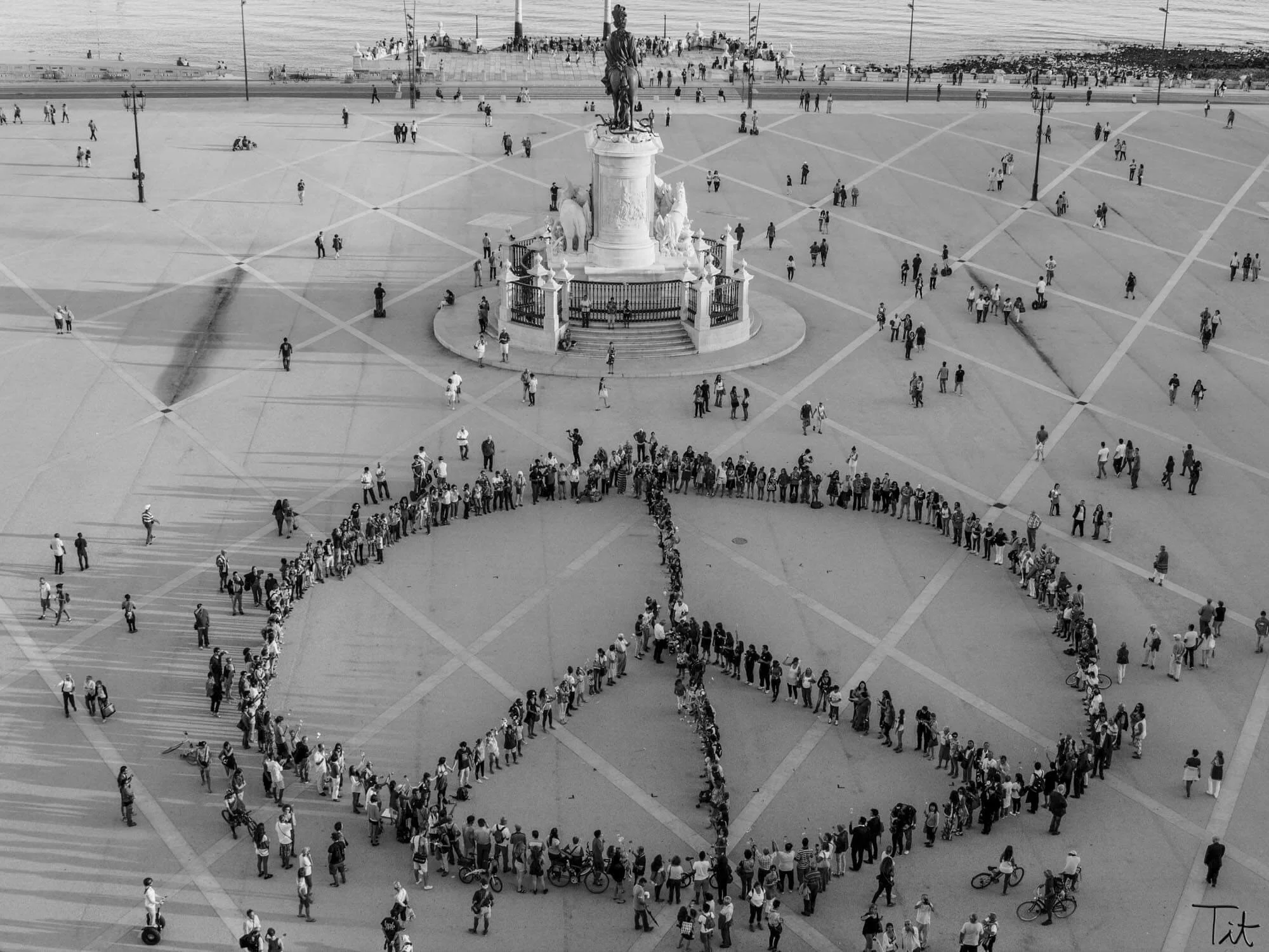 Black and white aerial photograph of a large open plaza with a statue at the center, surrounded by a circular fence and numerous people standing in long lines, forming peace symbols within the plaza.