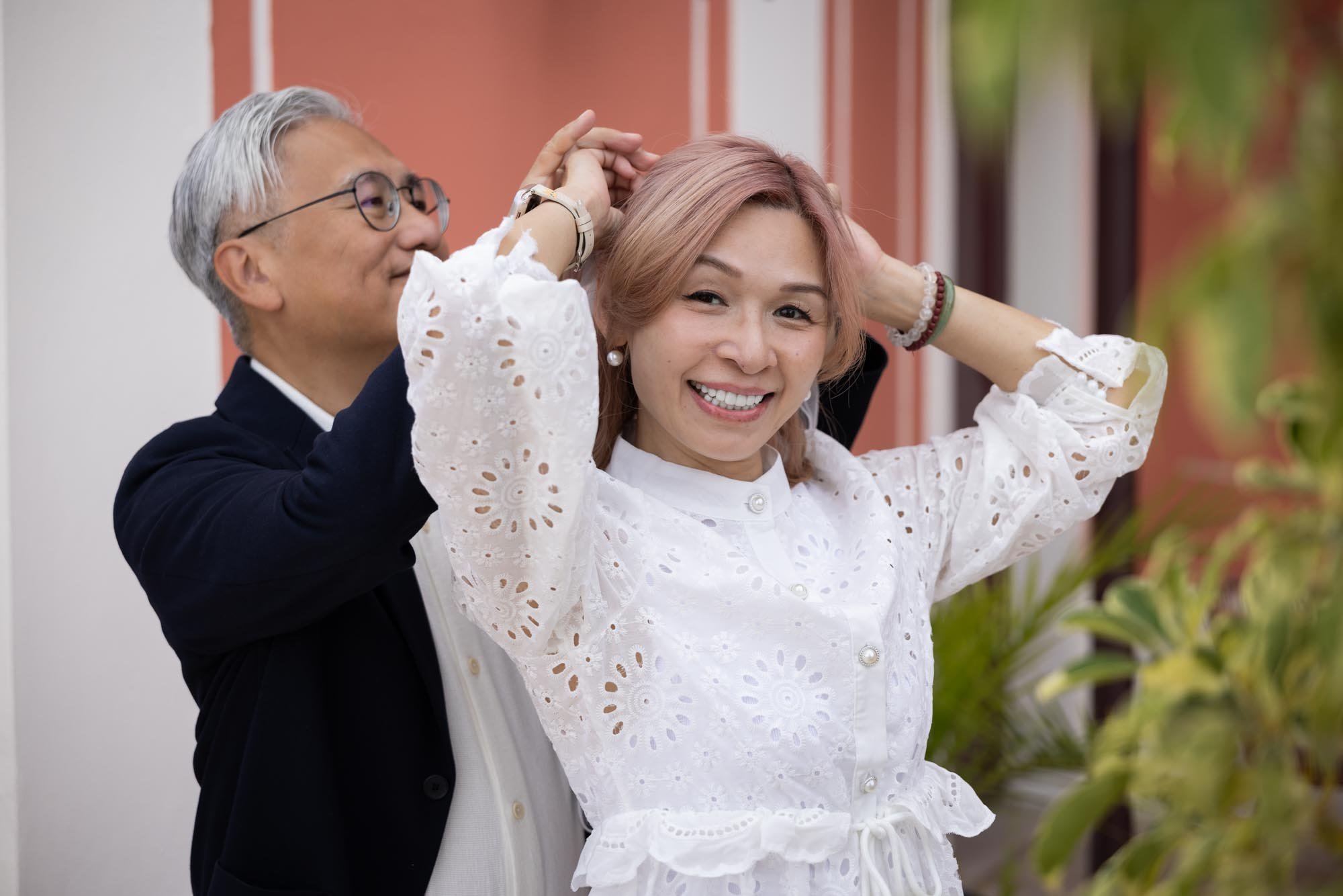 An older man with glasses helps a young woman with pink hair adjust her hair. The woman is smiling and wearing a white eyelet dress. They are outdoors in front of a building with red walls and green plants.