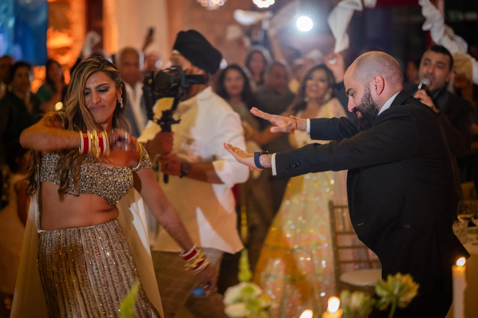 A woman in a sparkly dress is dancing with her eyes closed, while a man in a black suit with a beard is also dancing with his arms outstretched. The scene appears to be at a celebration or wedding with a lively, festive background and several onlooke