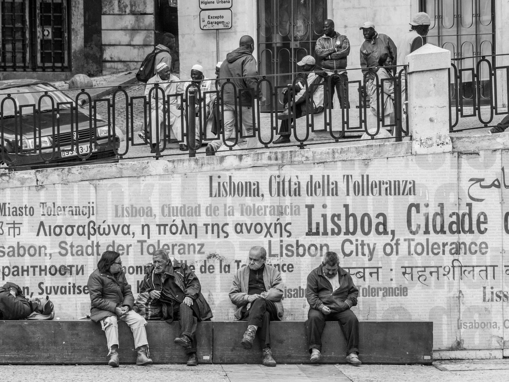 Black and white photo of people sitting and standing along a street in Lisbon, Portugal, with a large sign displaying the phrase 'Lisboa, Città della Tolleranza' in multiple languages.