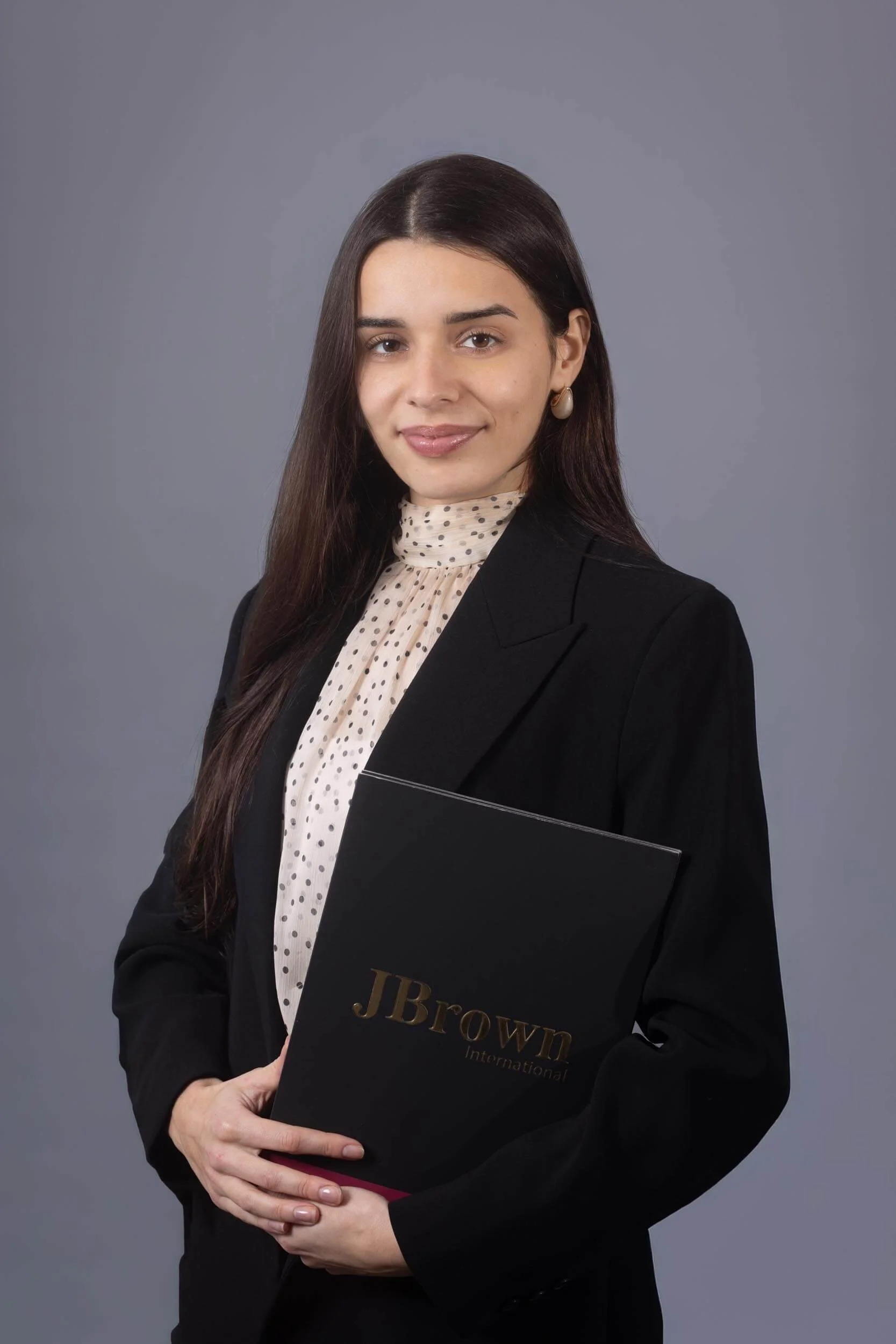 Professional woman holding a black folder labeled "JBrown International" against a gray background.