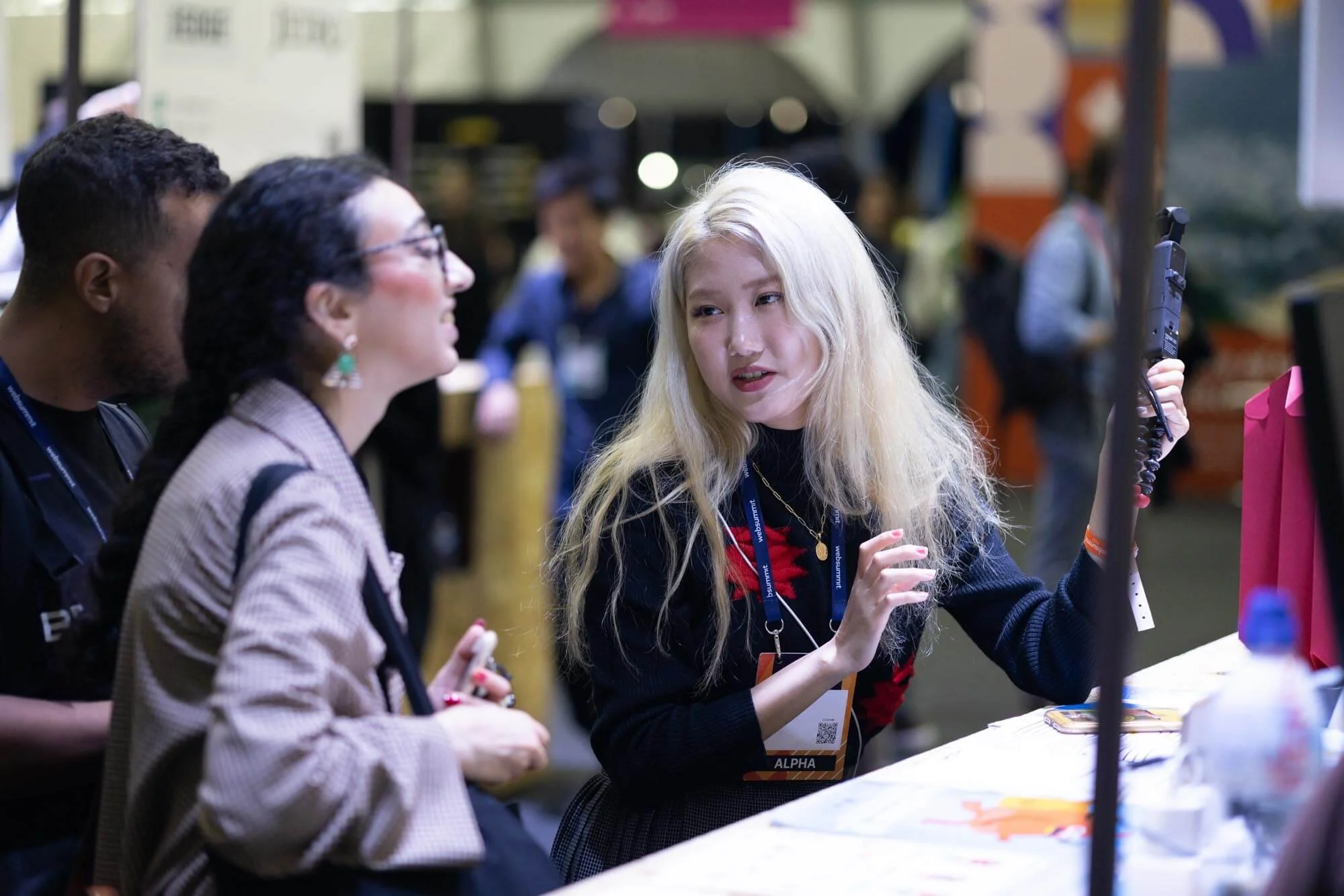 A woman with long blonde hair holding a gun and talking to two other women at a booth or table at an event.