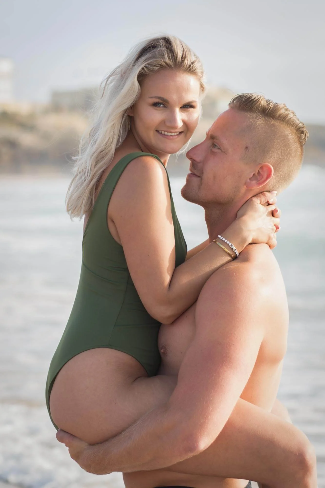 A man holding a woman in a green swimsuit on a beach with water and buildings in the background.