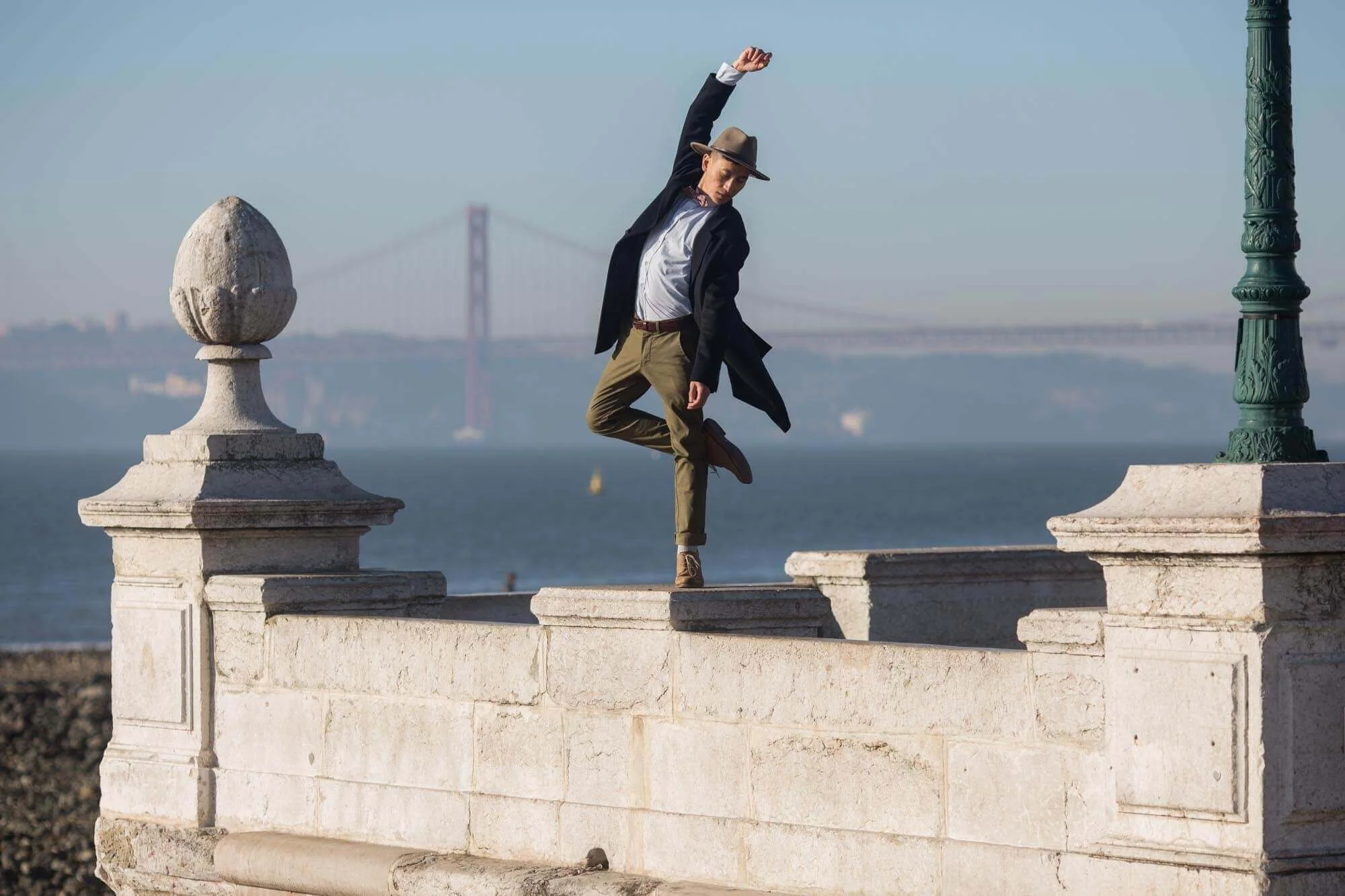 A man dressed in vintage-style clothing poses on a stone ledge by the water, with a bridge in the background.
