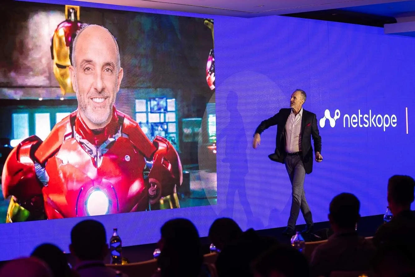 A man in a suit standing on stage next to a large screen that shows a person in an Iron Man costume. The stage backdrop displays the logo for Netskope.