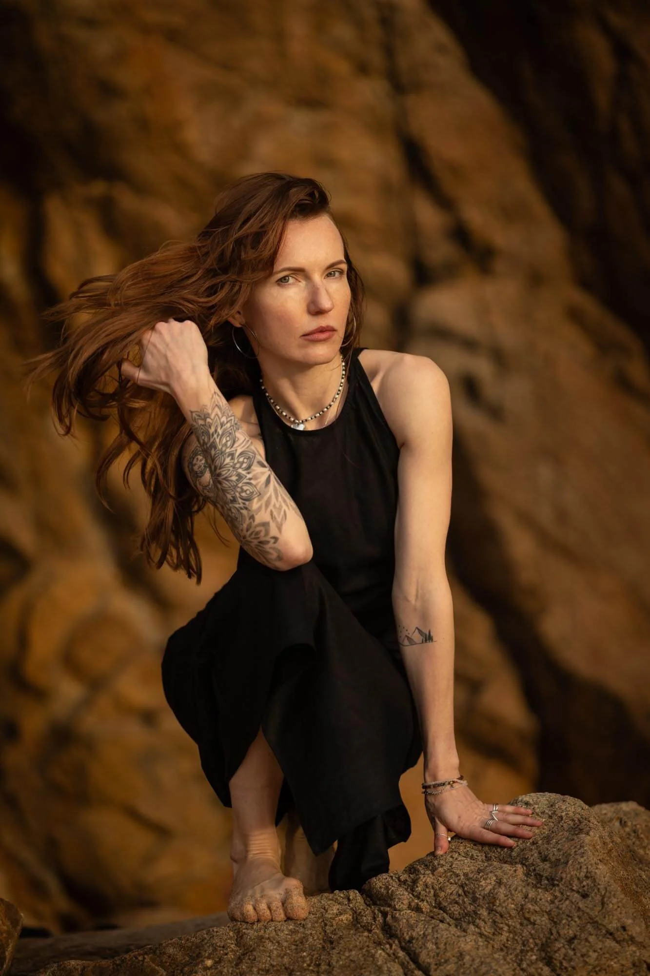 A woman with long red hair, tattoos on her arm and wrist, wearing a black sleeveless dress, crouching on rocks near a cliff with a serious expression, holding her hair back with her left hand.