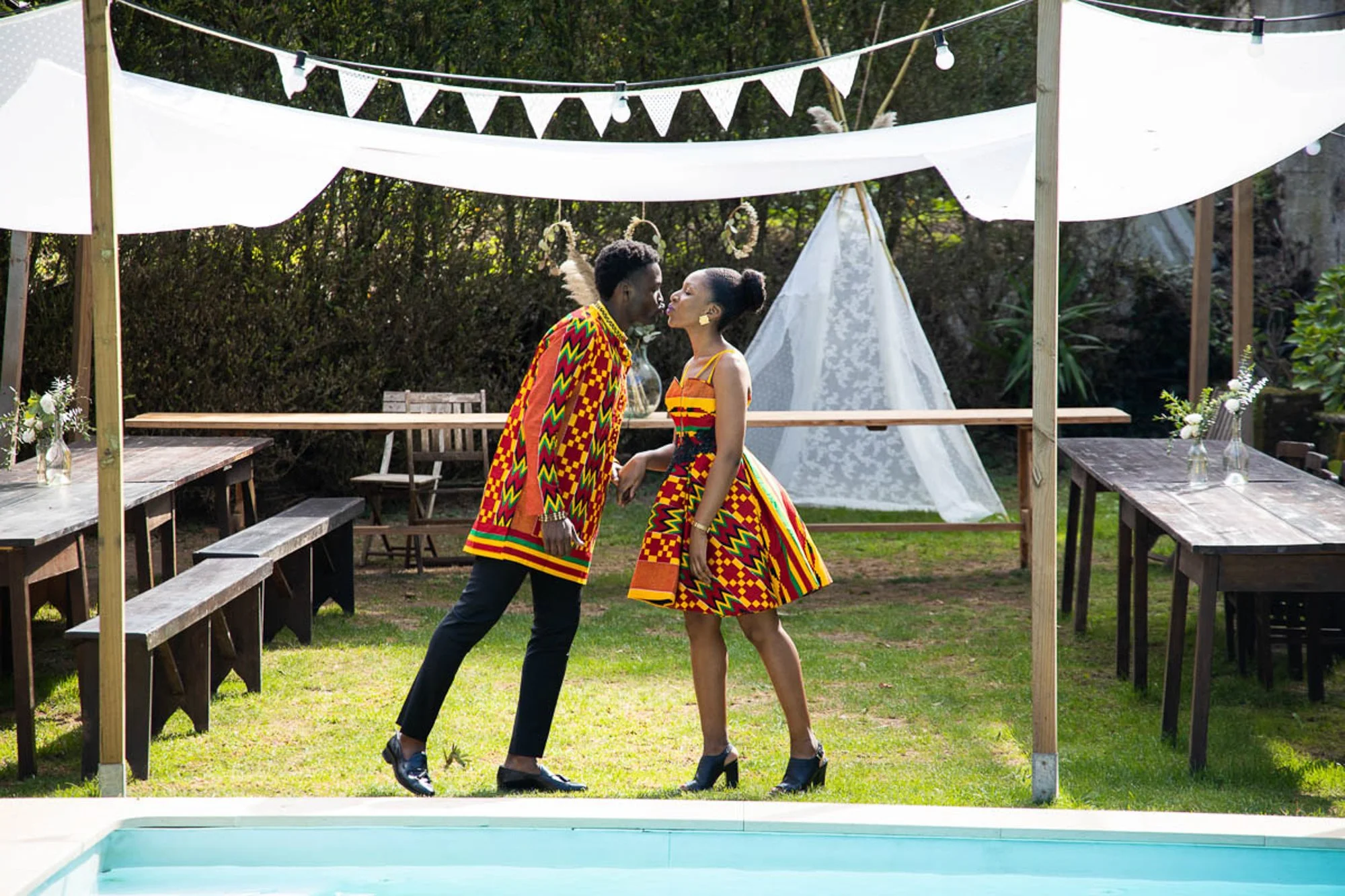 A couple dressed in bright, colorful African prints dancing by a swimming pool, with outdoor tables decorated with vases of flowers and a white canopy overhead.