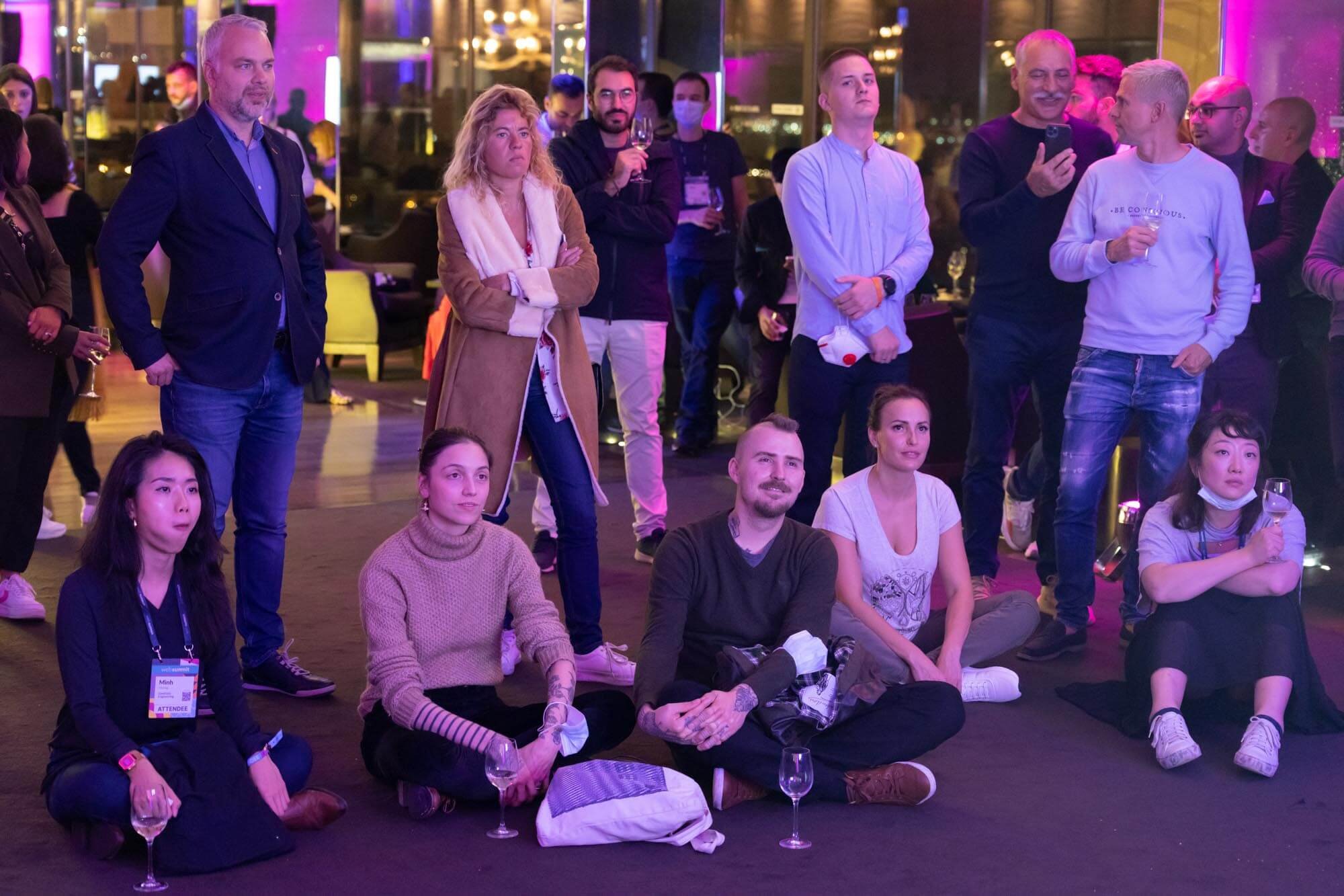 A group of people, some sitting on the floor and others standing behind, attending a social event in an indoor venue with purple lighting, some holding wine glasses, and a few wearing attendee badges.