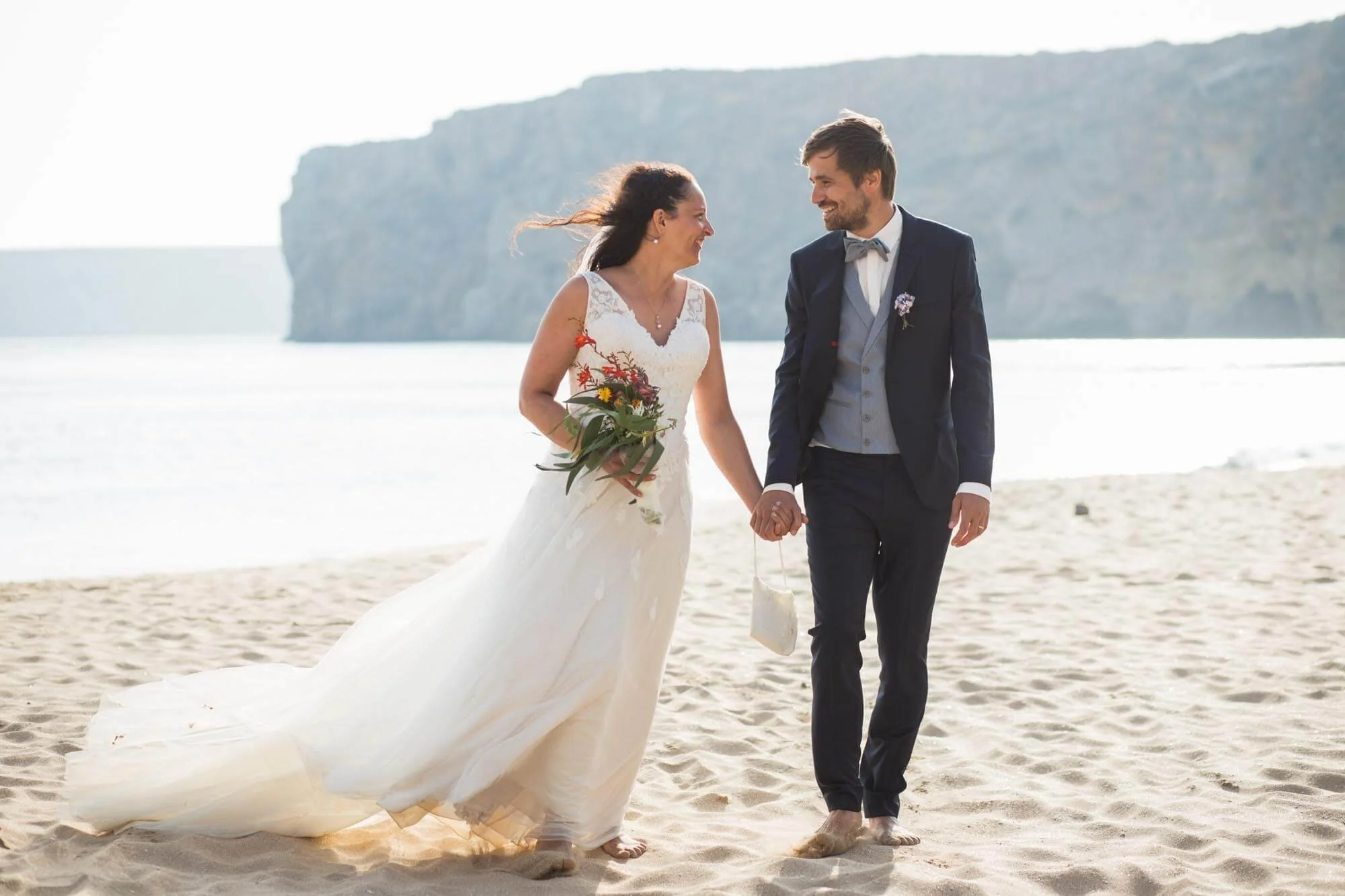 A bride and groom walking hand in hand on a sandy beach, smiling at each other, with a large cliff and the ocean in the background.