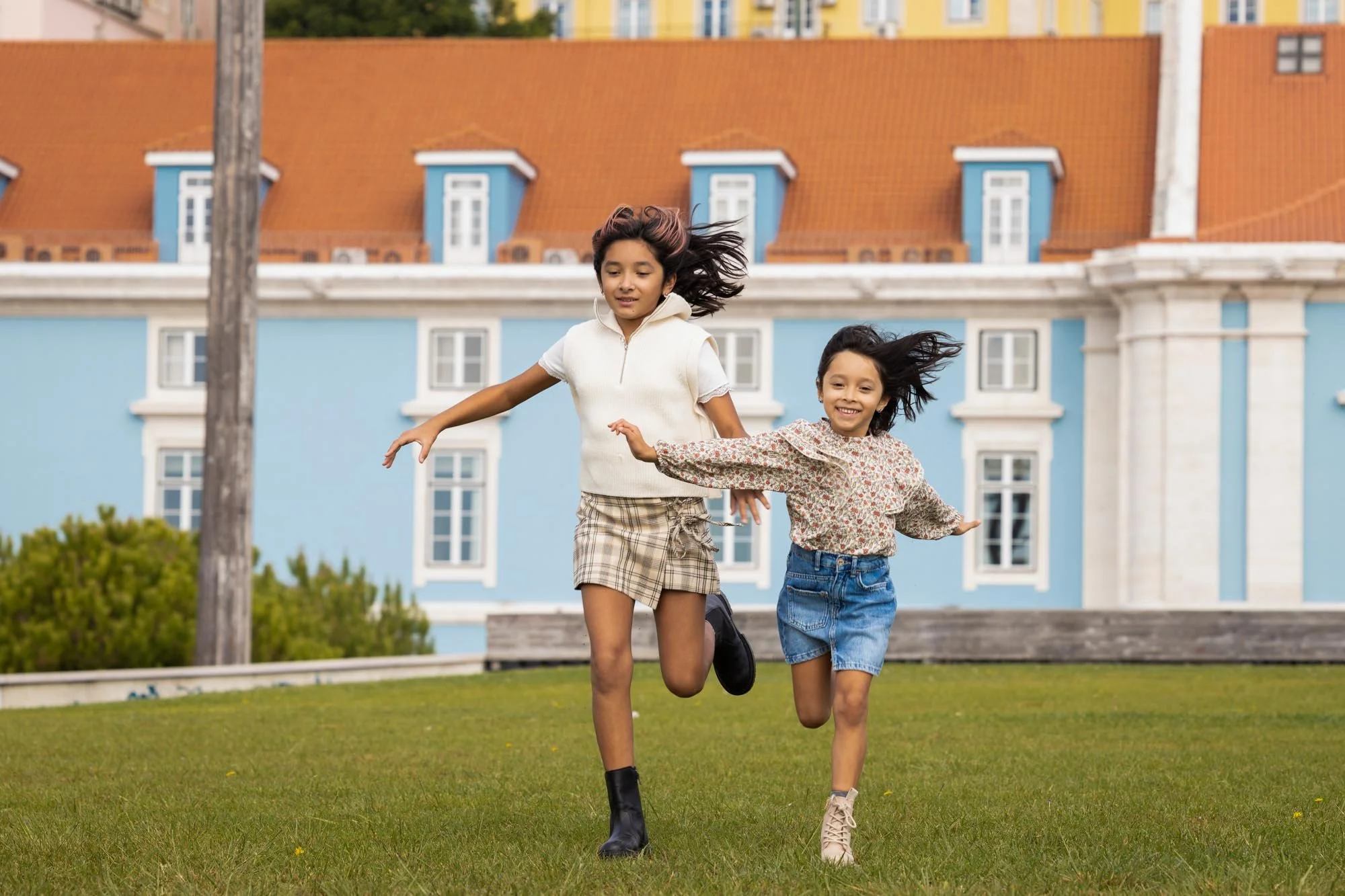 Two young girls are running on a grassy lawn in front of a light blue building with a red roof.