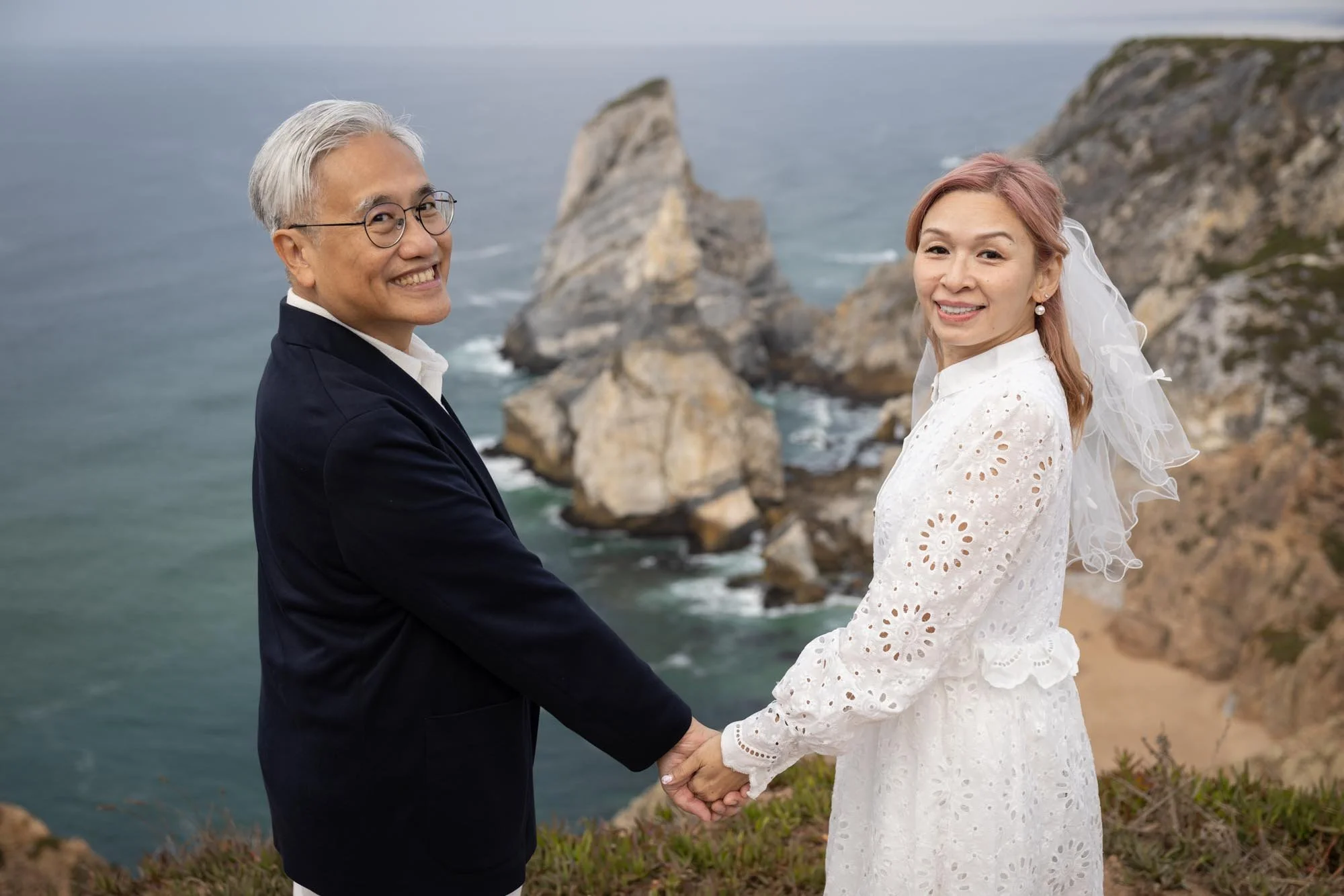 A couple holding hands in front of a rocky coastal landscape, dressed in formal attire, with the man in a dark suit and glasses, and the woman in a white dress and veil, smiling at the camera.