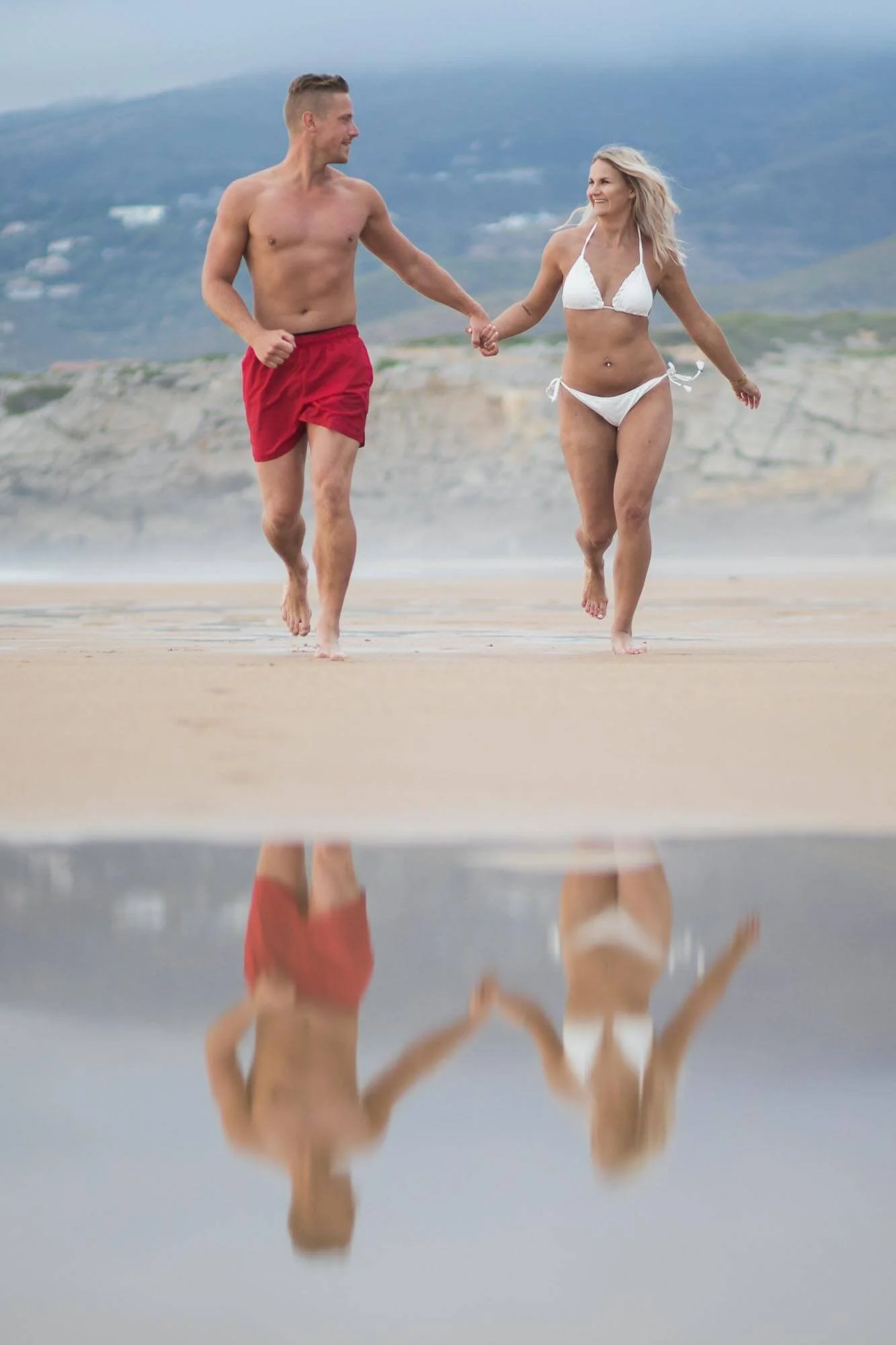 A man and woman in swimwear walking on the beach holding hands, with their reflection visible in a shallow puddle or wet sand.