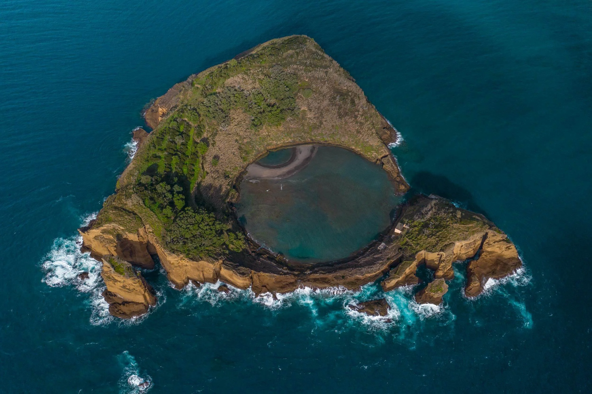 Aerial view of a small island surrounded by blue ocean with a large natural volcanic crater pool filled with water, lush green vegetation, rocky shores, and waves crashing against the edges.