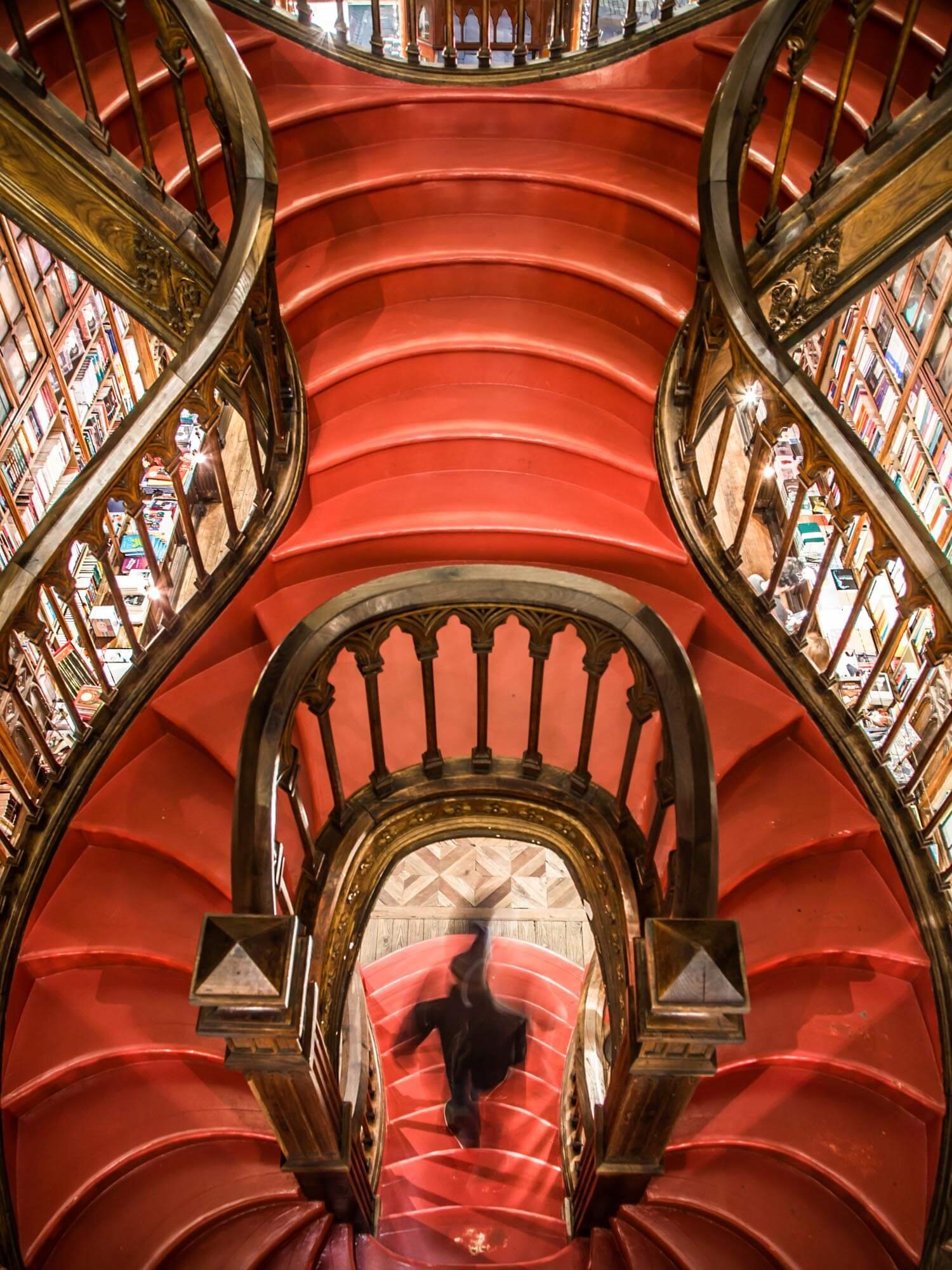 Overhead view of a red velvet carpeted spiral staircase with ornate wooden and gold trim, in a library with bookshelves filled with books visible on the sides.
