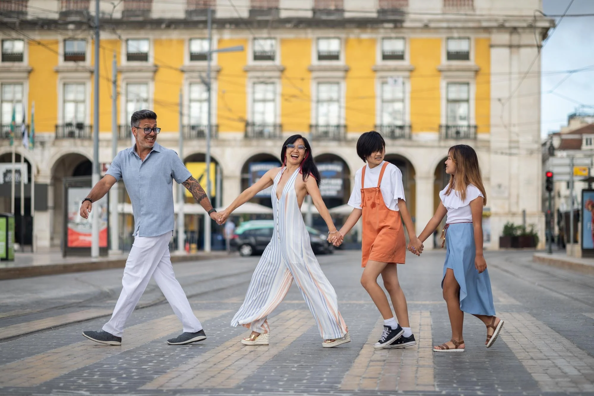A diverse group of five people holding hands and walking across a city street crosswalk, smiling and enjoying a sunny day with a yellow building in the background.