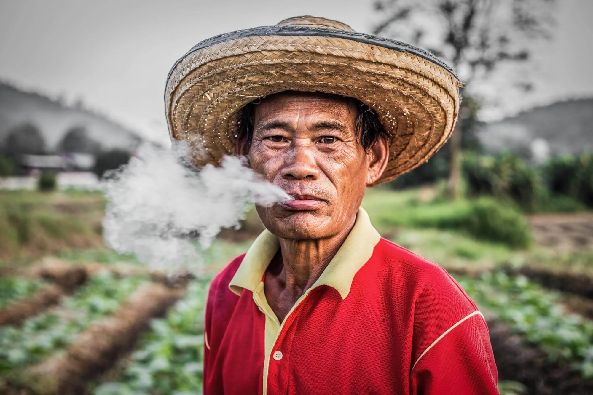 An older man in a red shirt and a large straw hat, smoking with smoke visible near his mouth, standing outdoors with blurred greenery and farmland in the background.