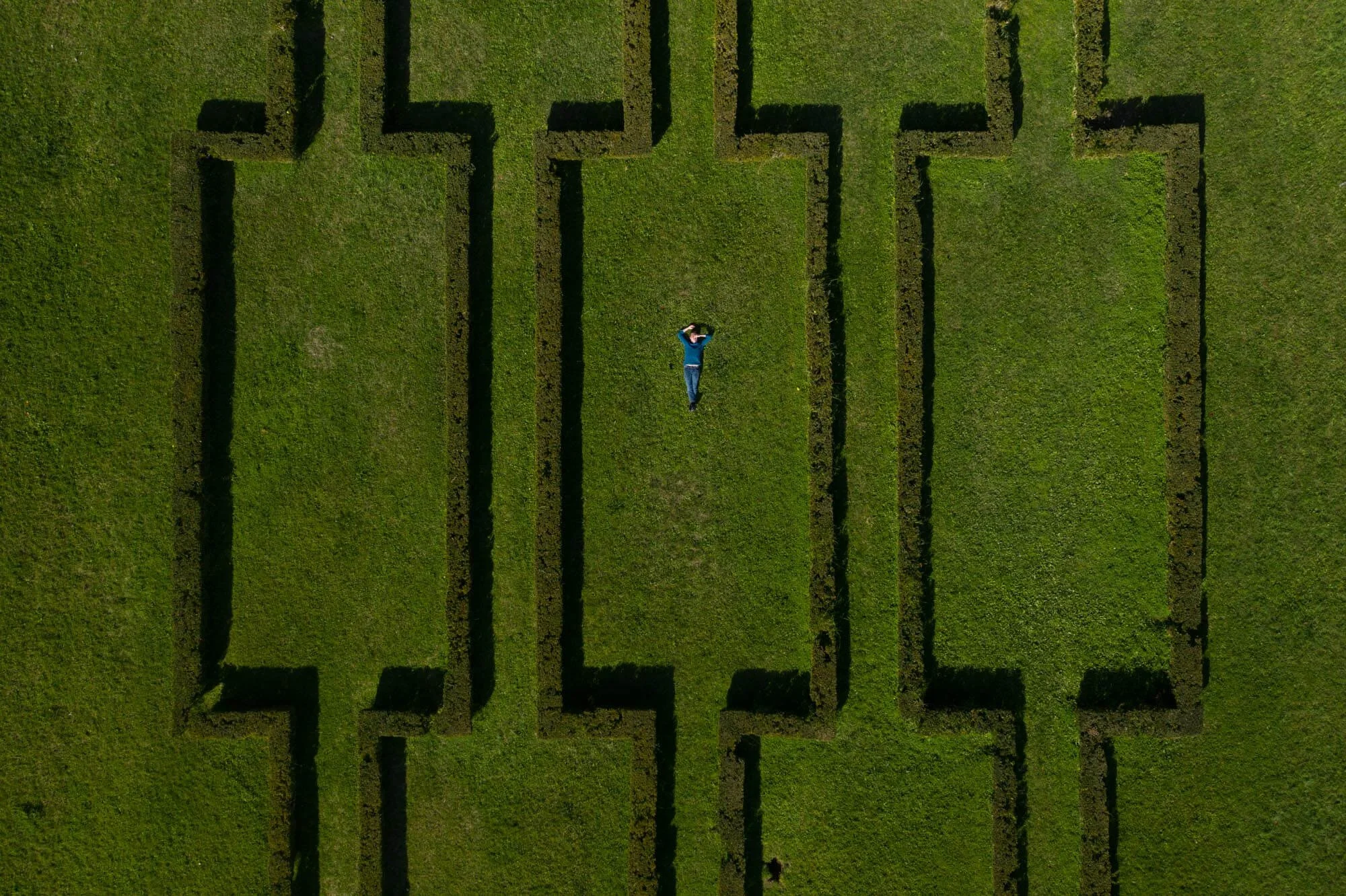 A person lying on a green grassy field within a maze with tall hedge walls viewed from above.