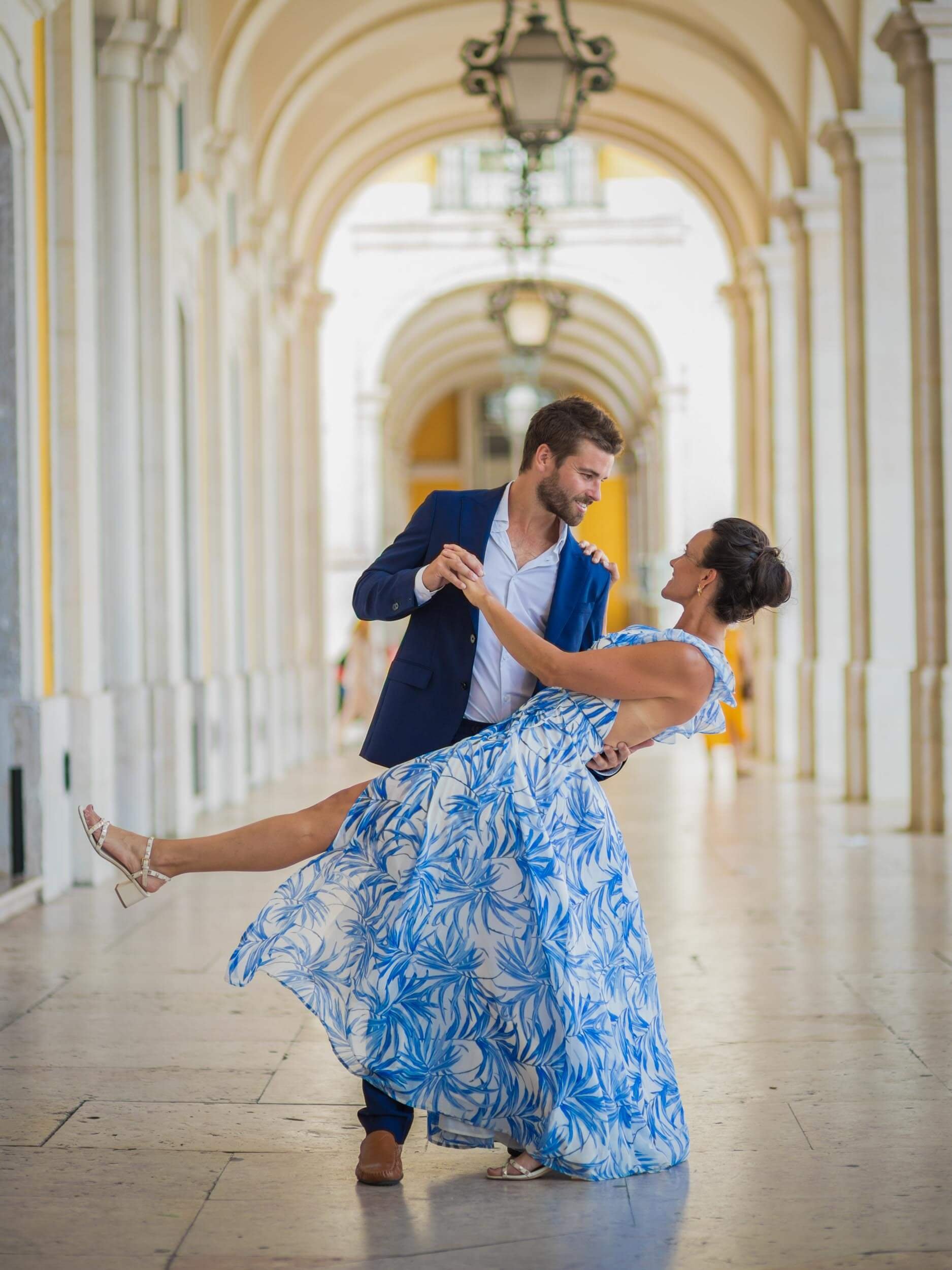 A man in a blue suit is dancing with a woman in a blue and white patterned dress, in an elegant covered walkway with arches and hanging lanterns.