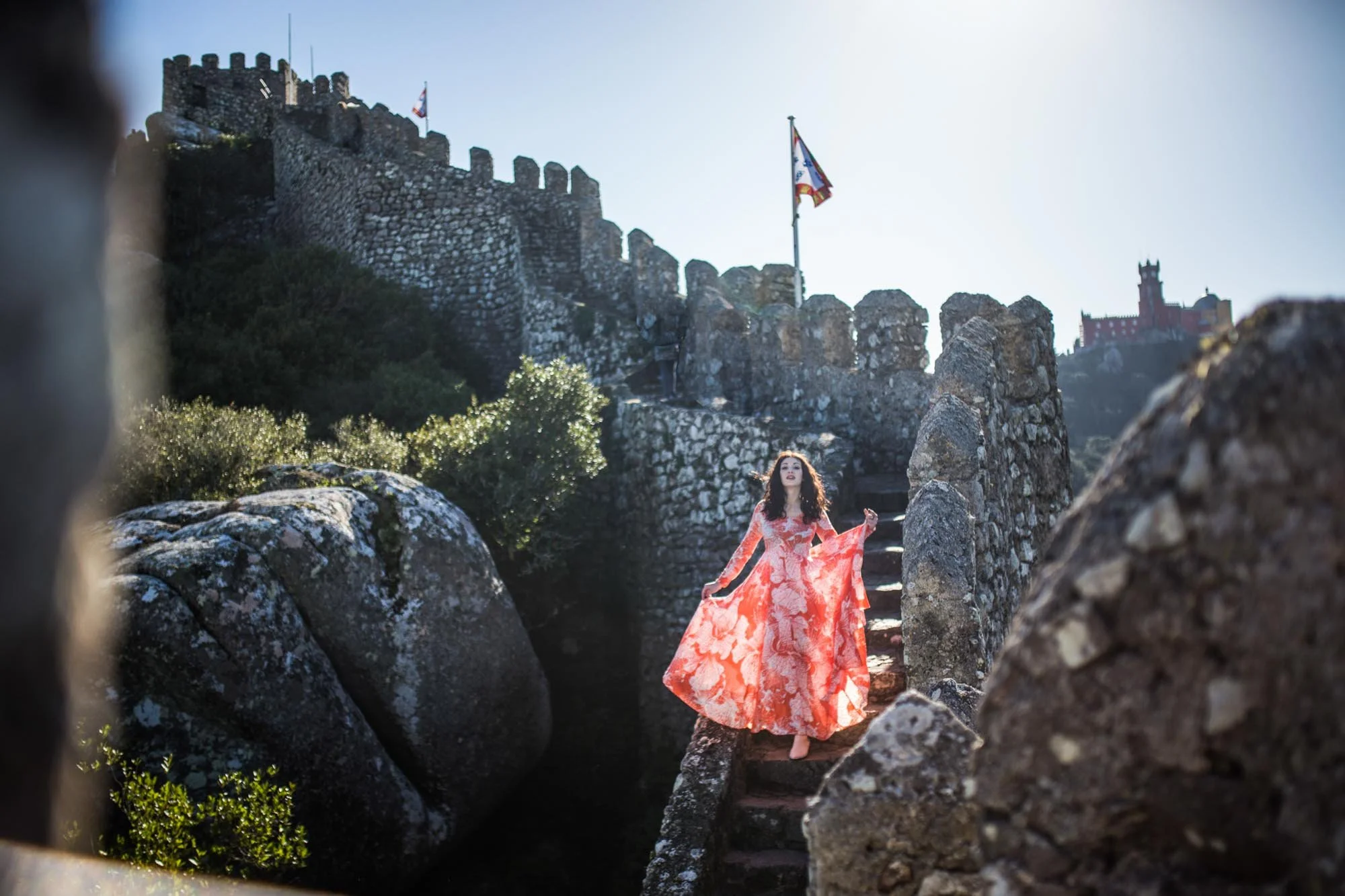 Woman in a flowing orange floral dress standing on stone stairs between large rocks, with castle walls and flags in the background.