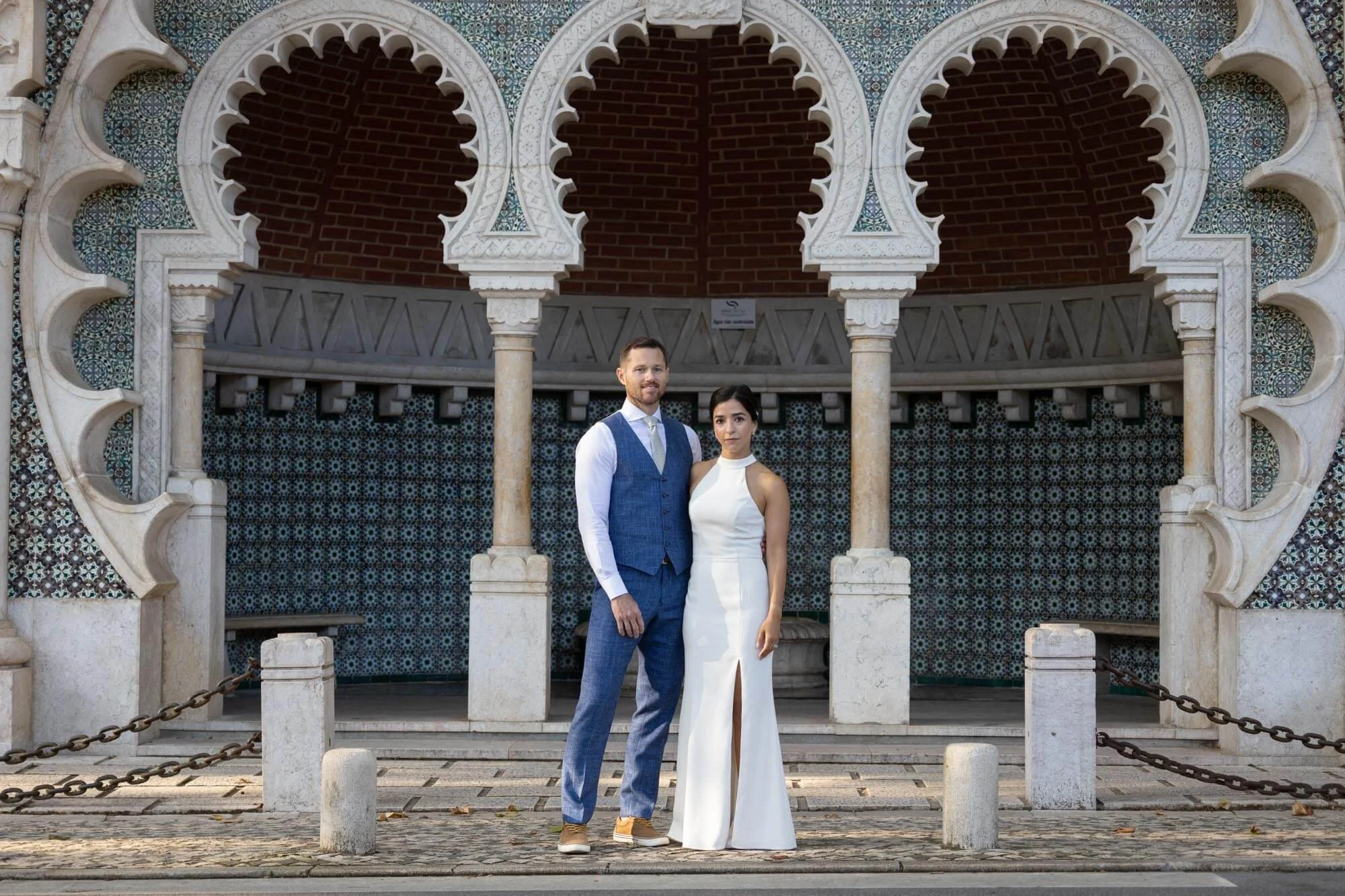 A couple dressed in formal attire, the man in a blue vest and pants with a white shirt and tie, and the woman in a white sleeveless gown with a slit, standing in front of an ornate, decorative archway with intricate patterns and a tiled wall.