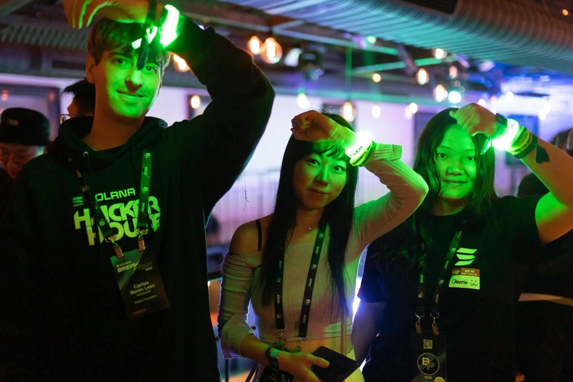 Three young people at an indoor event, wearing black t-shirts and wristbands, making a salute gesture. They are under string lights with colorful glow, and are wearing event badges.