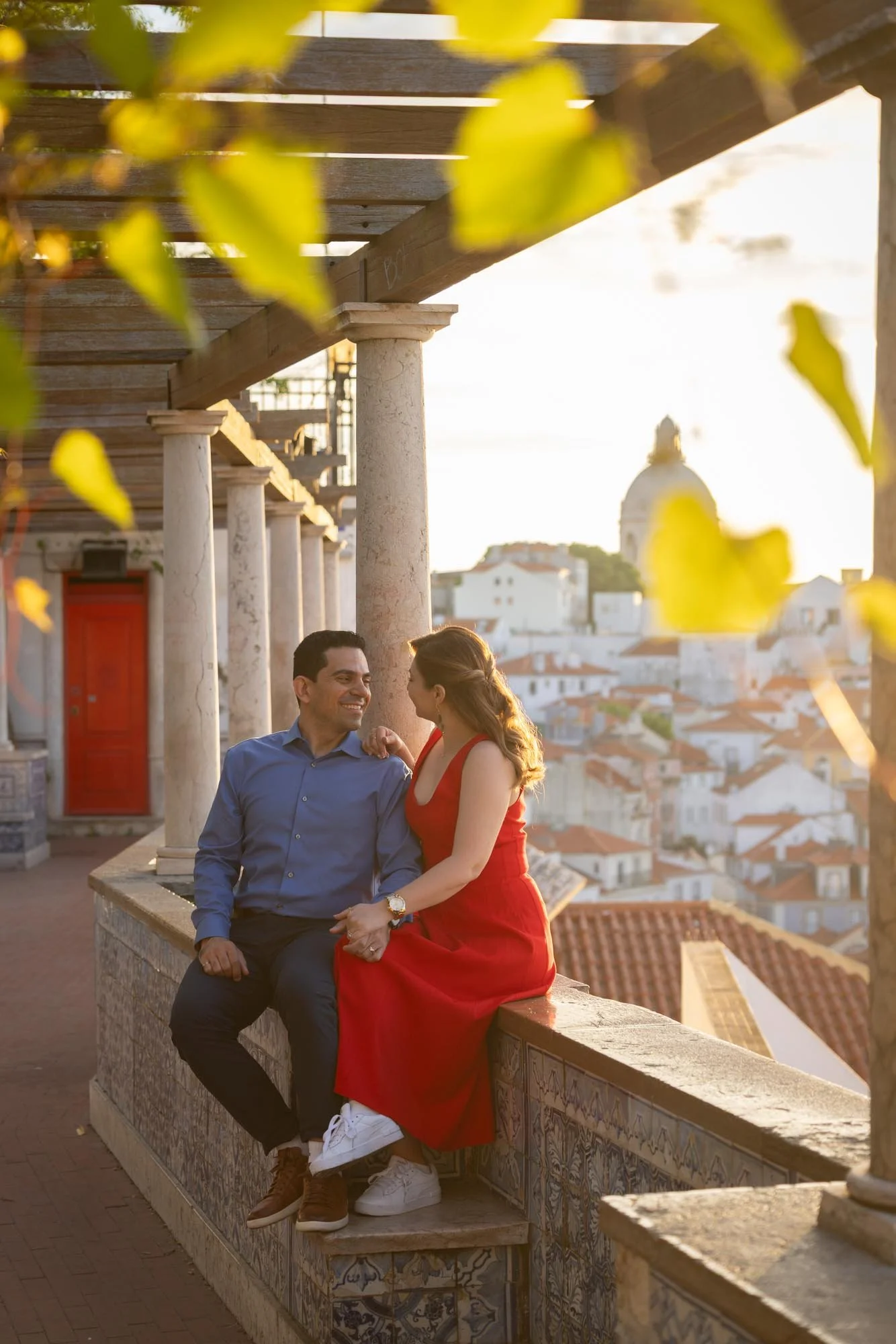 A man and woman sitting on a decorative tiled ledge, enjoying each other's company in an outdoor setting with historic architecture and a cityscape in the background during sunset.