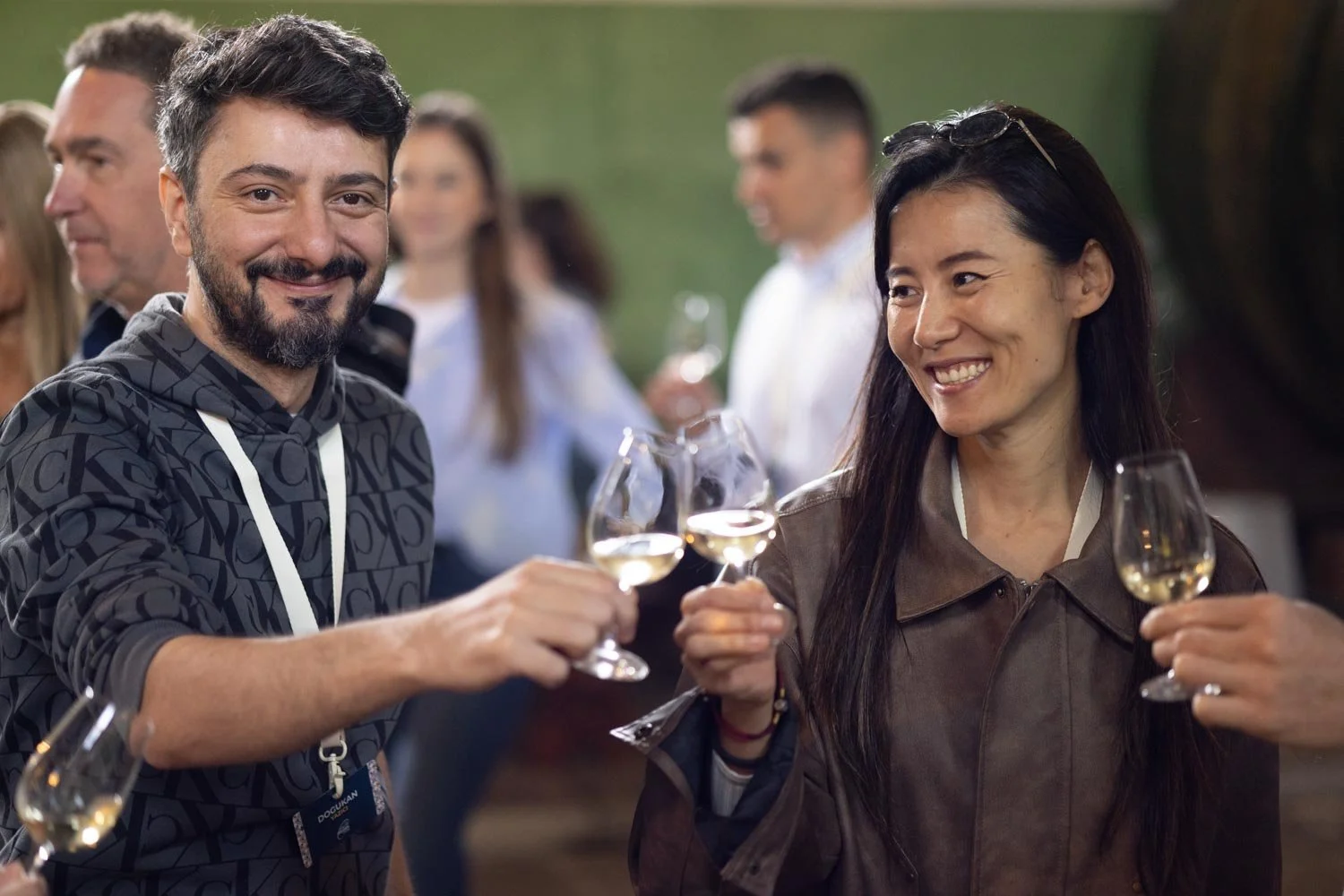 A group of people at a social gathering outdoors, raising glasses of white wine for a toast, with smiling faces and blurred people in the background.
