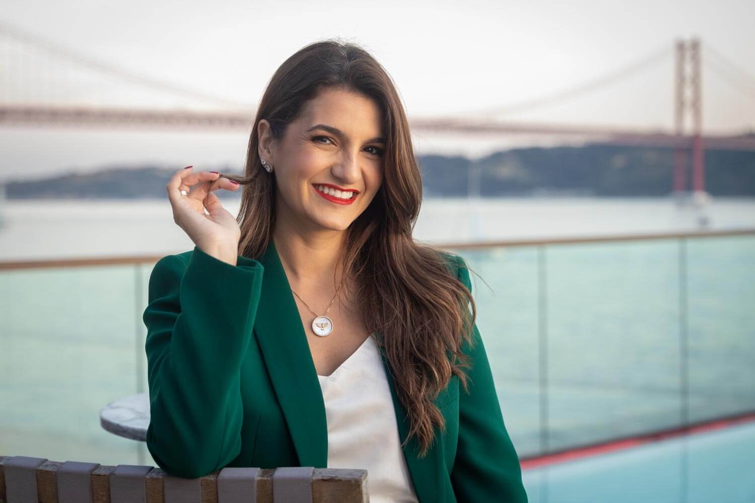 A smiling woman in a green blazer and white top sits outdoors near water with a bridge in the background.