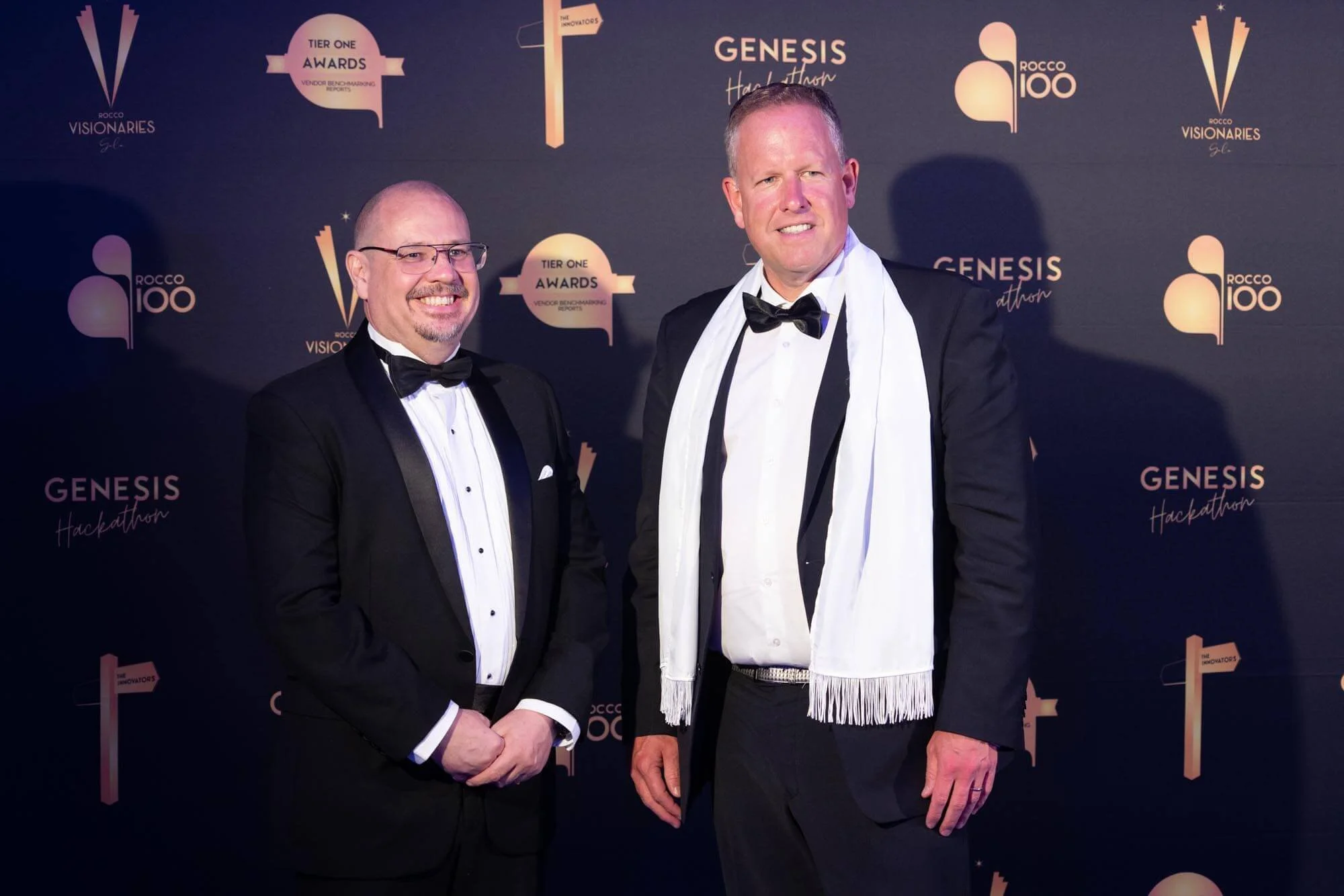 Two men in tuxedos standing on a black carpet at a formal event, with a backdrop displaying logos and text such as 'Rocco 100', 'Genesis Hackathon', and 'Tier One Awards'.