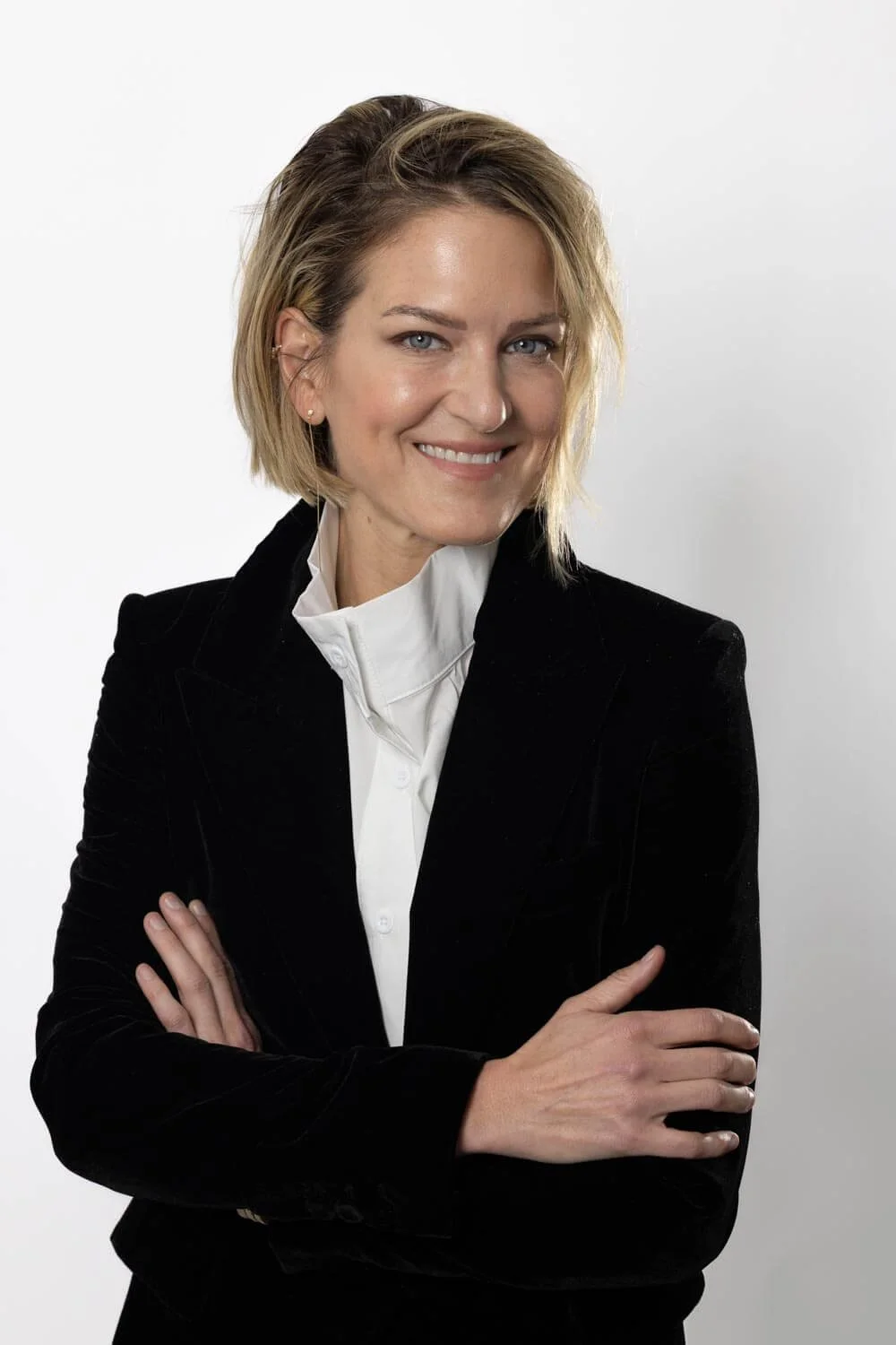 Woman with blond hair, wearing a black blazer and white blouse, smiling with arms crossed against a white background.