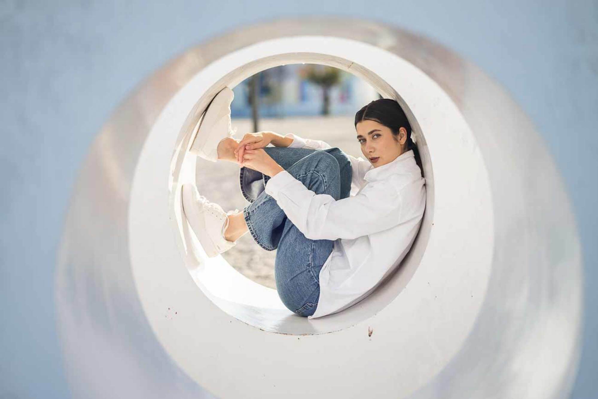 A woman sitting inside a large white circular structure, possibly a park sculpture, wearing a white shirt and blue jeans, looking at the camera.