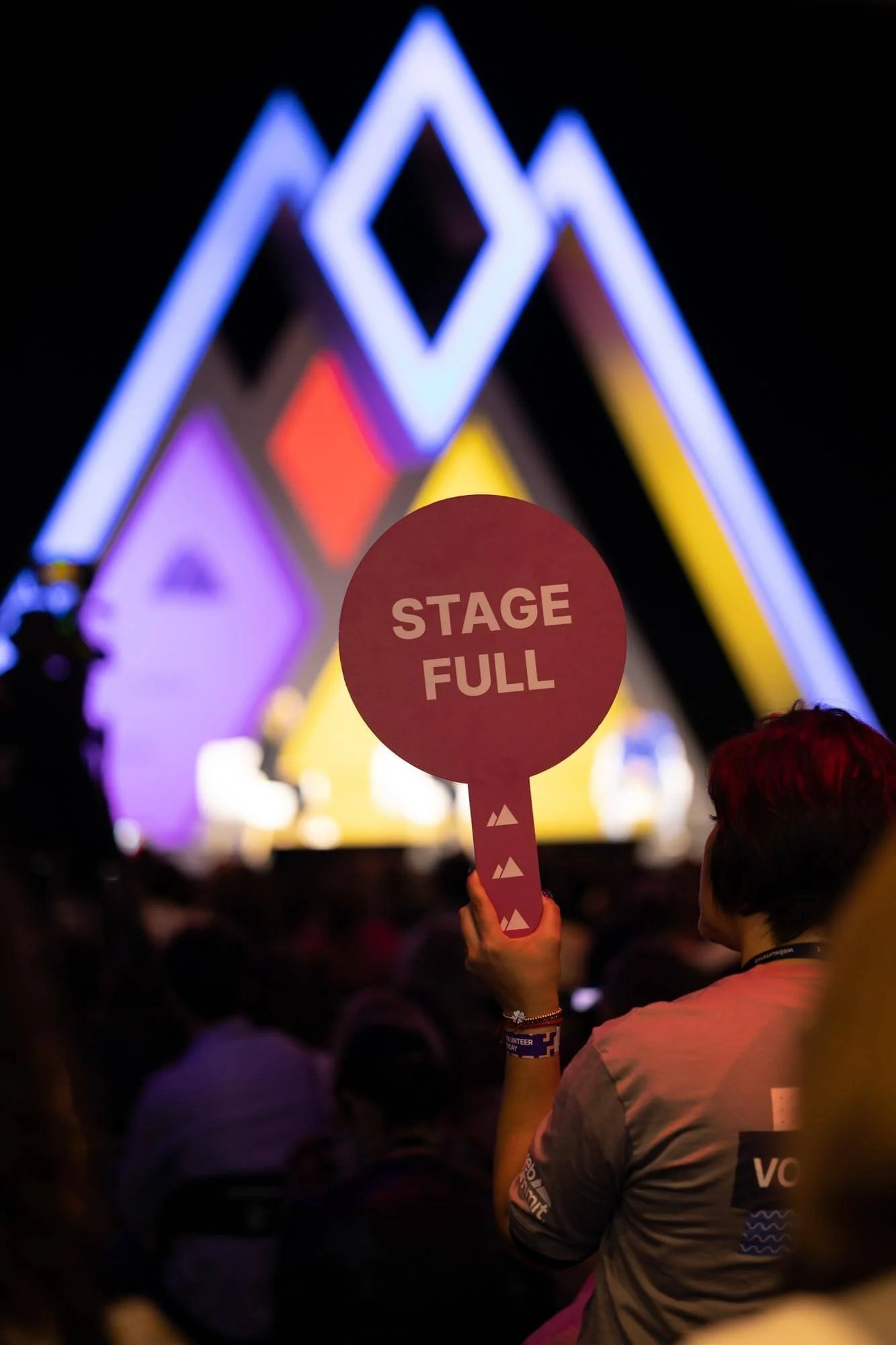 A person holding a pink circular sign that reads 'STAGE FULL' during a concert or event, with a colorful illuminated stage in the background and a dark crowd surrounding them.