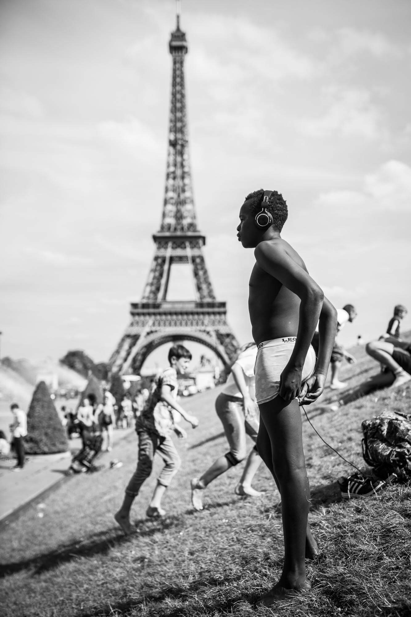 A black and white photo of children playing in front of the Eiffel Tower, with one boy in the foreground wearing headphones and underwear.