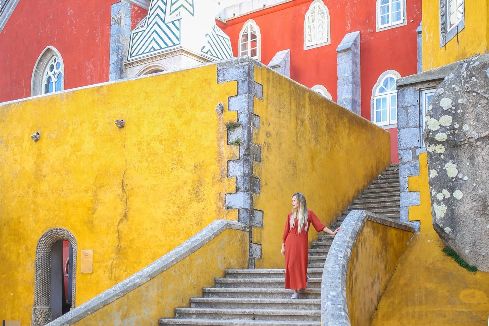 A woman in a long red dress standing on a staircase with a yellow wall and colorful buildings with arched windows in the background.