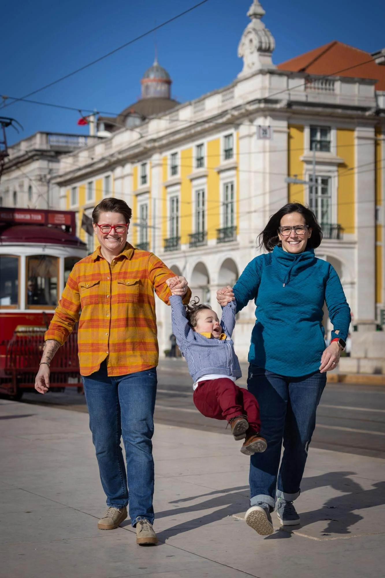 Two women are walking outdoors on a sunny day, holding a young girl between them by her hands. The women are smiling and appear happy. The background features a colorful building and a tram, indicating an urban setting.