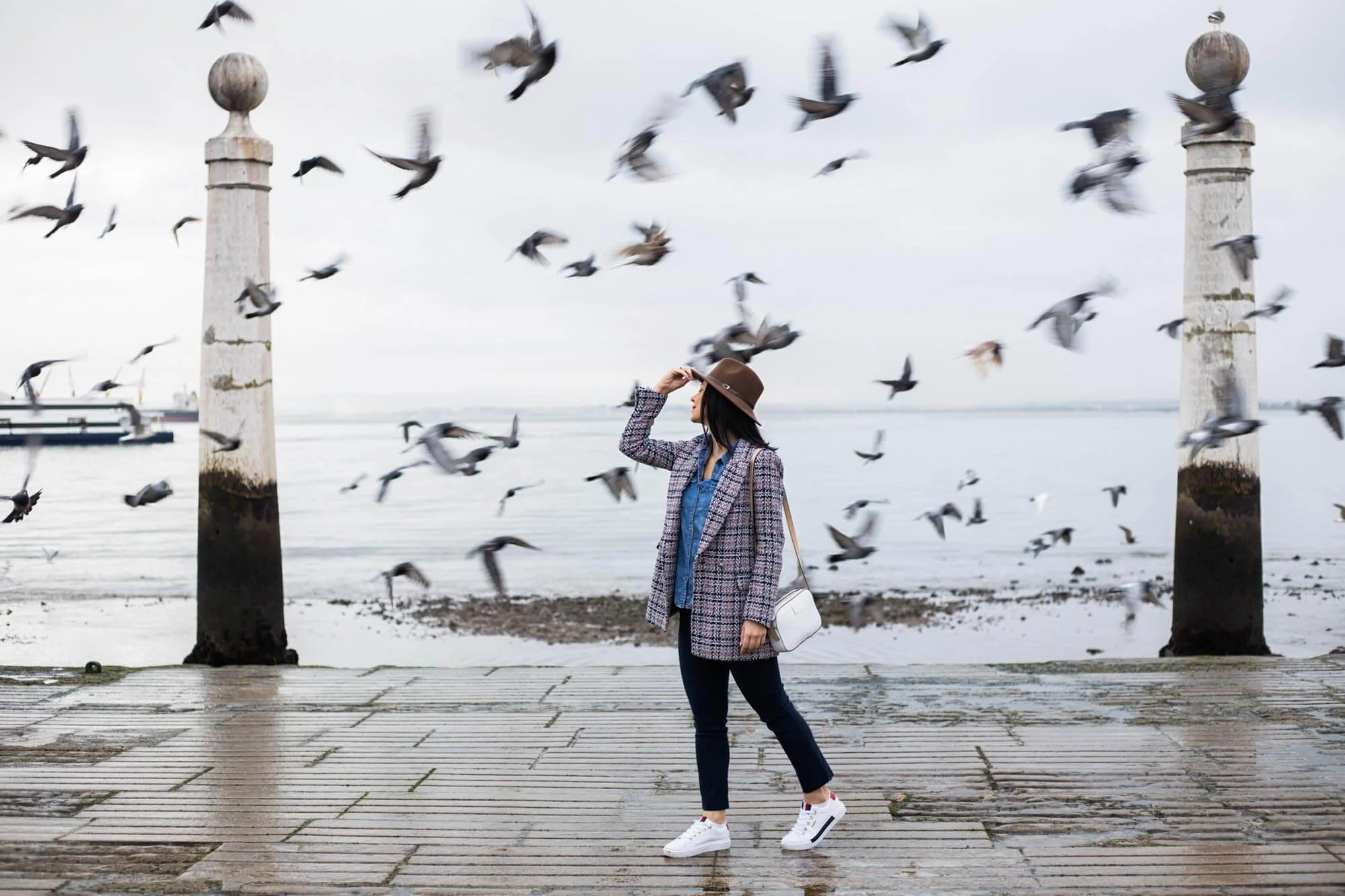 A woman in a blue shirt, plaid coat, dark jeans, white sneakers, and a brown hat stands on a dock near water, surrounded by flying birds.