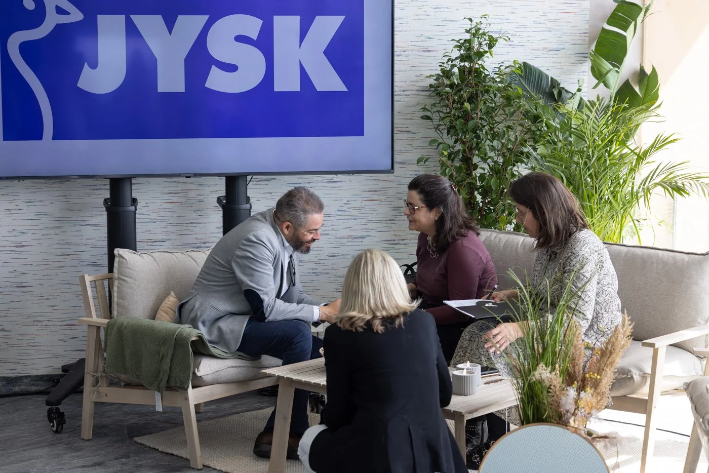 A group of five people engaged in a discussion in an indoor setting, with a large screen displaying the JYSK logo behind them and surrounded by green plants.