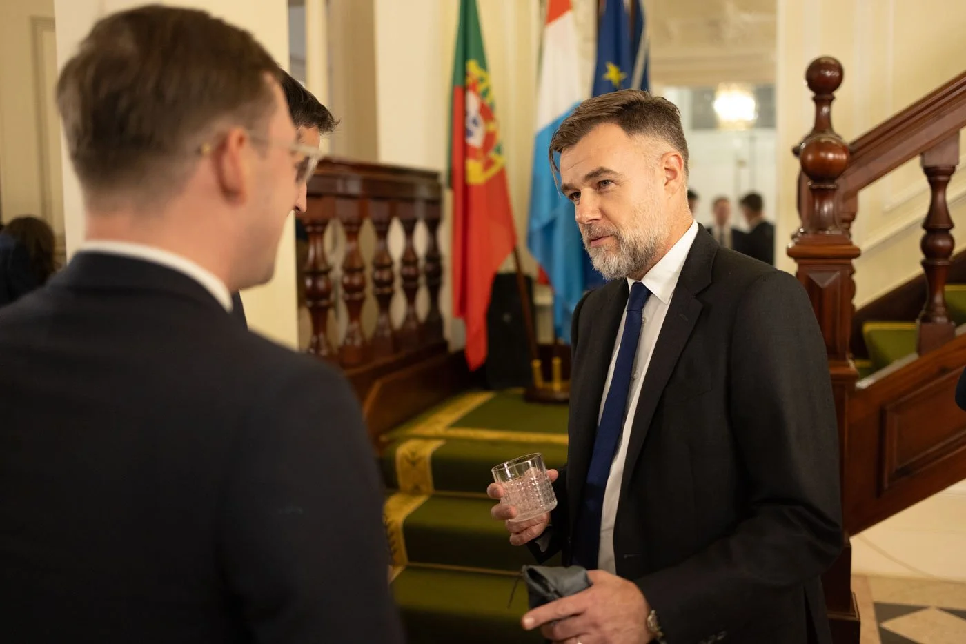 Two men in suits having a conversation in a formal setting, with flags in the background.