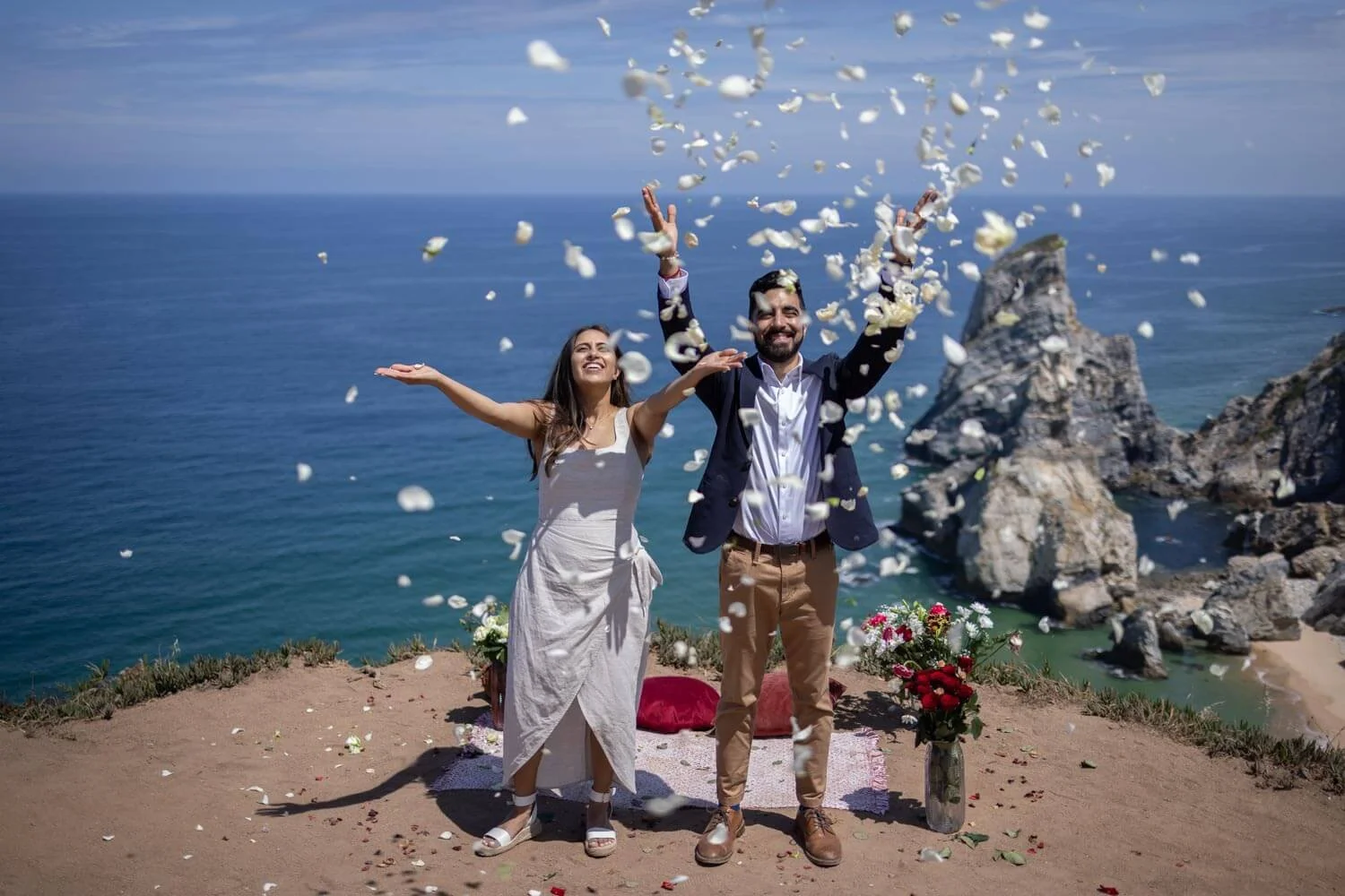 A couple celebrating on a cliff near the ocean, throwing flower petals into the air, with rocks and the sea in the background, under a partly cloudy sky.