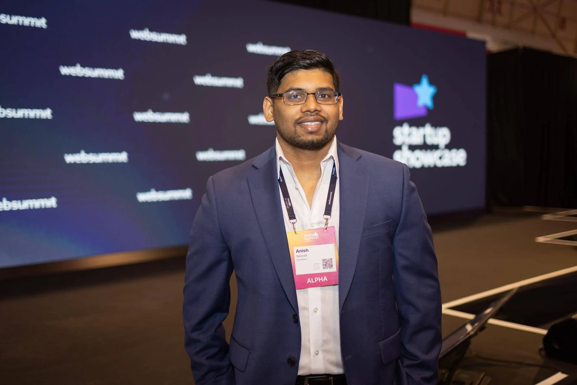 A man in a dark blue suit and white shirt standing in front of a stage with a large screen displaying the 'Startup Showcase' logo at a conference or event.