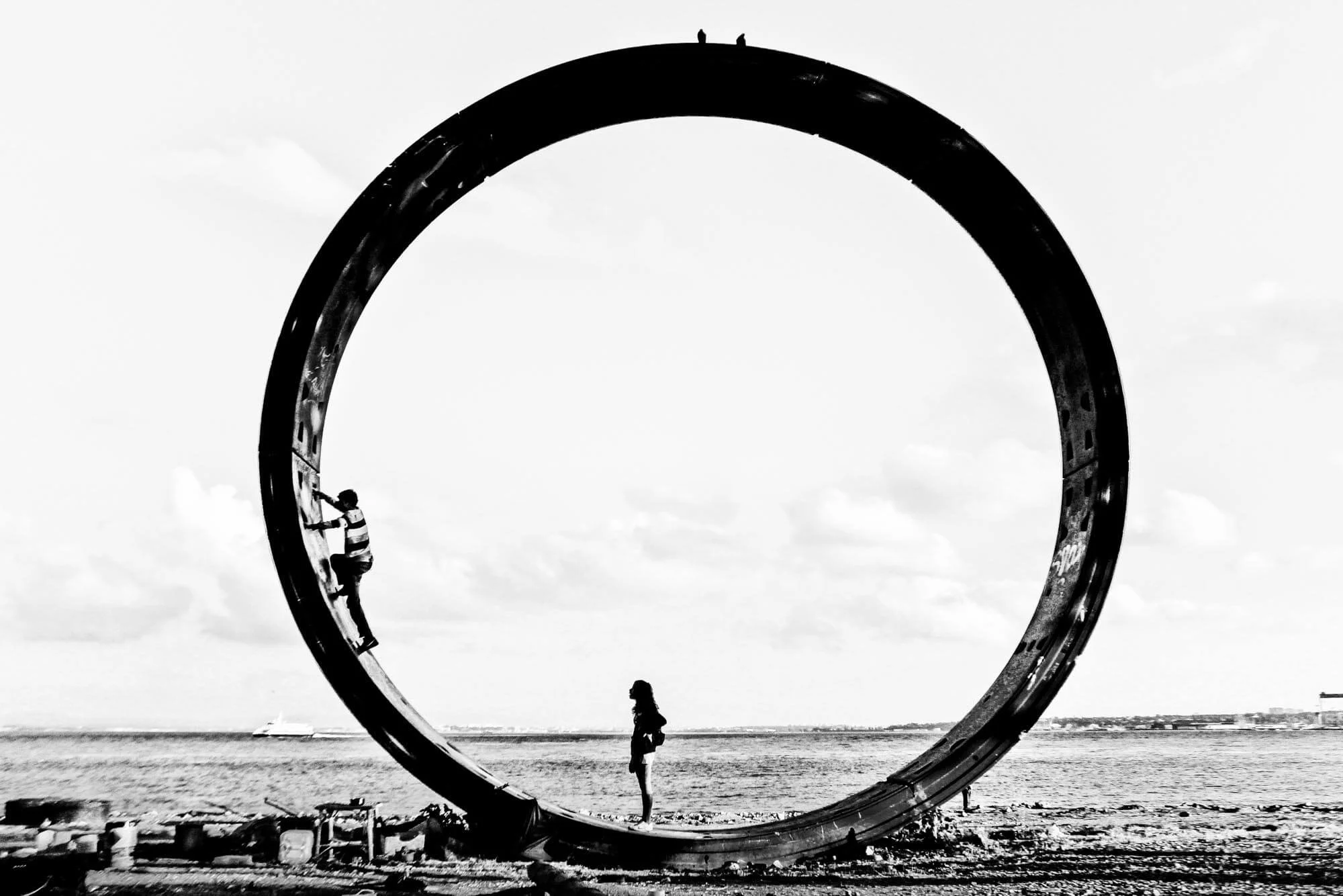 A black and white photo of a large circular sculpture on a beach with four people interacting with it. One person is climbing on the sculpture, while another stands inside the circle, and two people are standing on top of it.