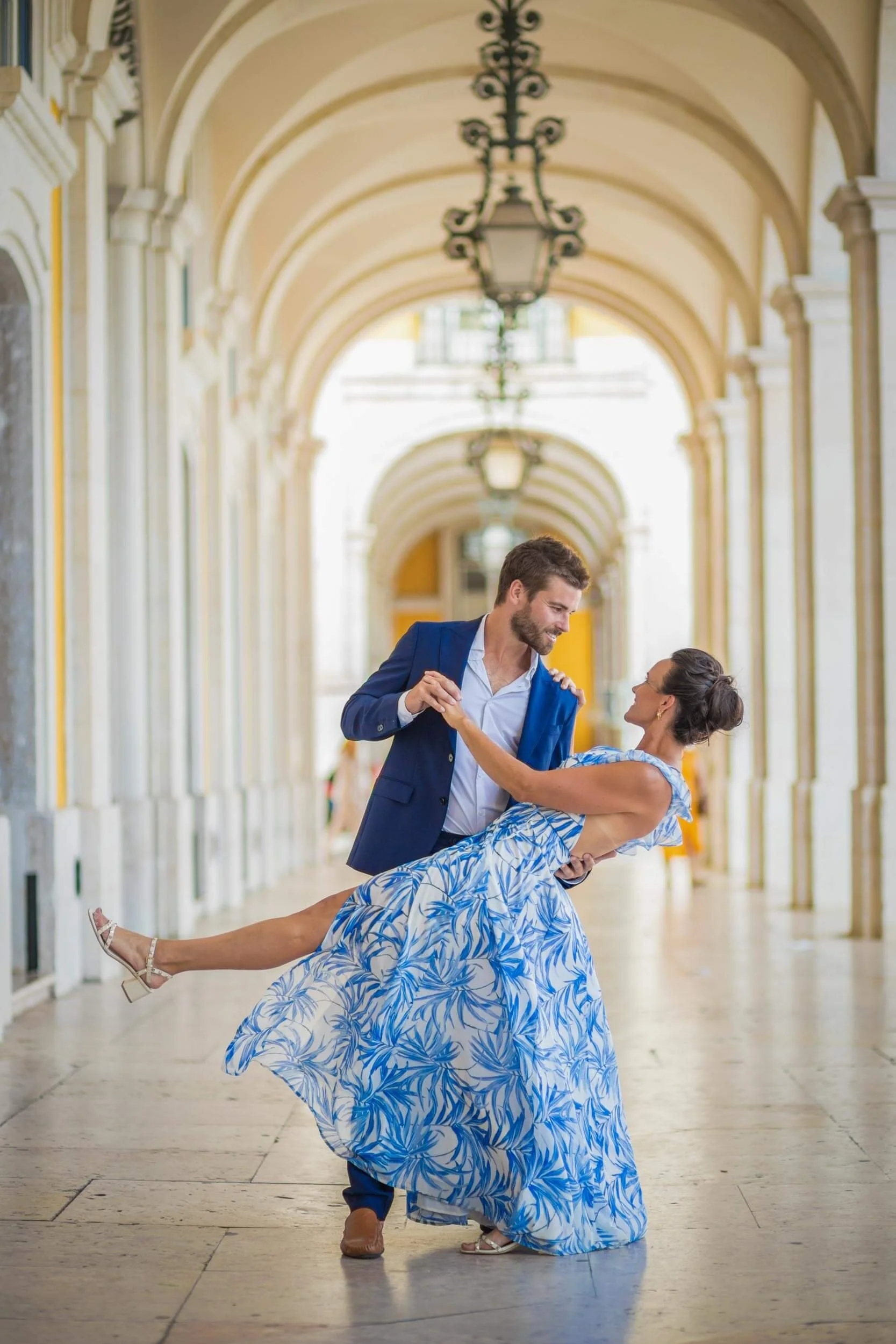 A Lisbon couple photoshoot with a man in a blue suit dancing with a woman in a blue and white patterned dress in an elegant, arched corridor with hanging lanterns.